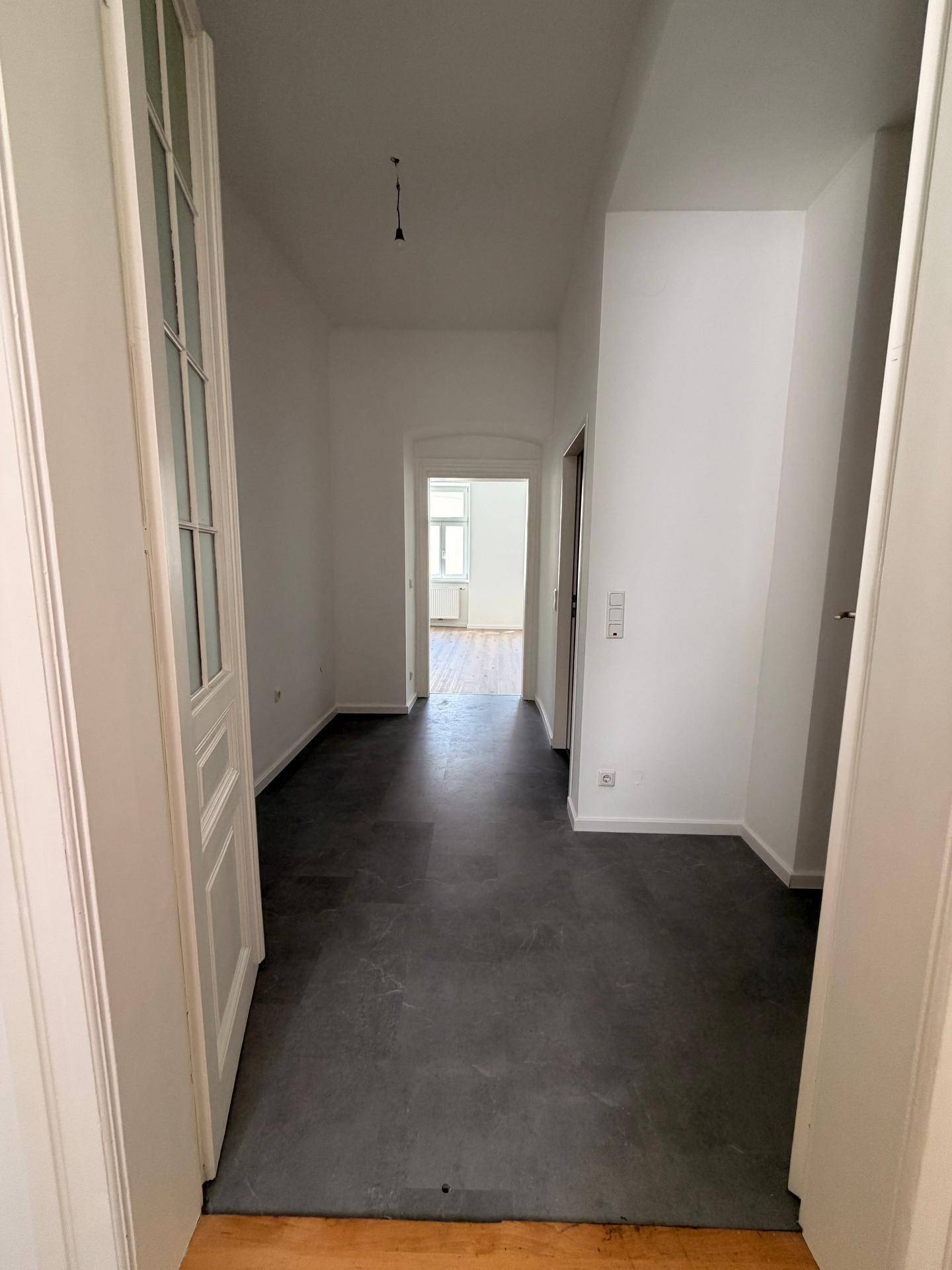 Heller Flur mit dunklem Fliesenboden und Durchgang zu weiteren Räumen mit Holzboden.