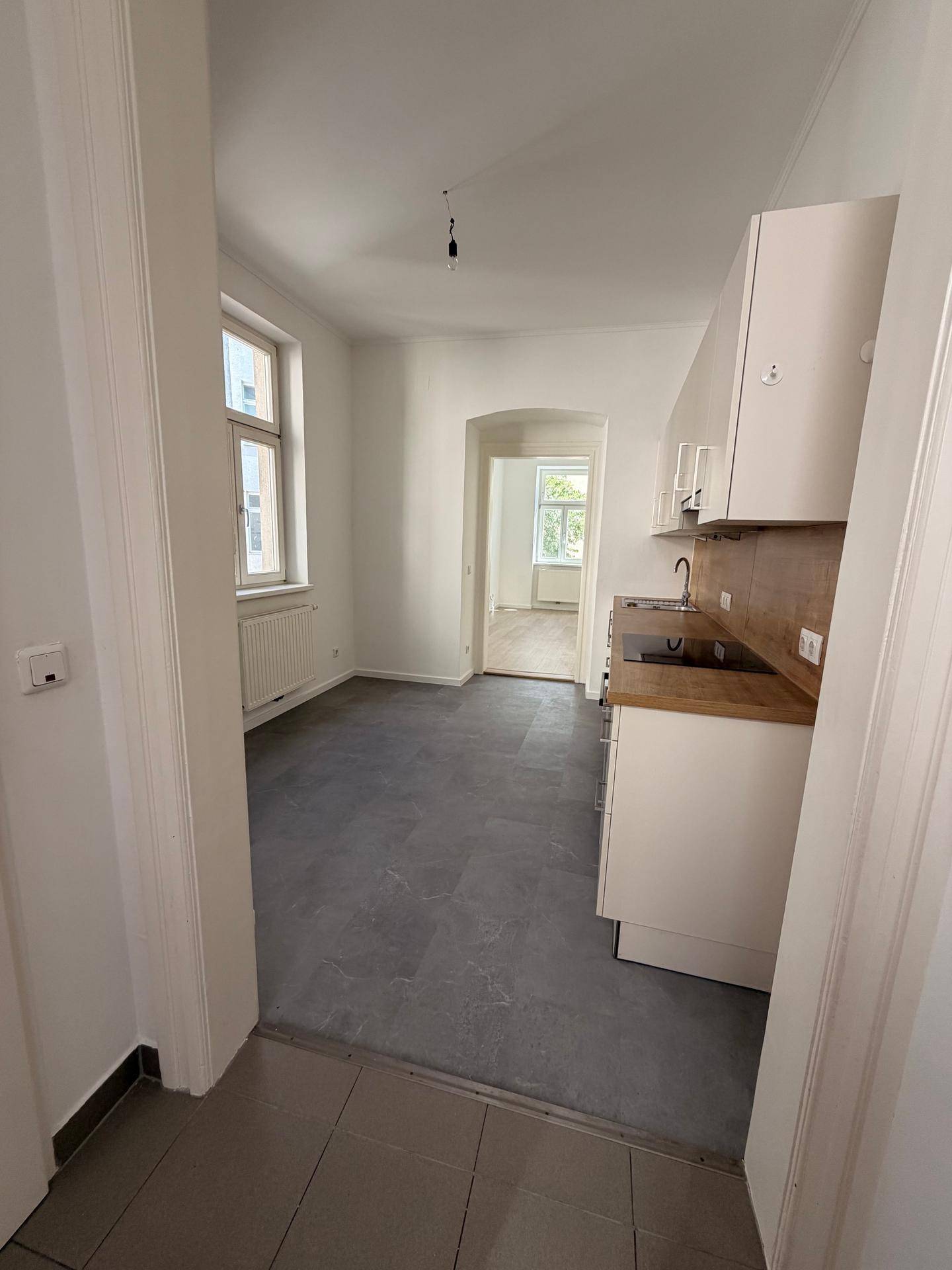 Offener Wohnbereich mit kleiner Küchenzeile, Fliesenboden und Durchgang zu einem weiteren Zimmer.