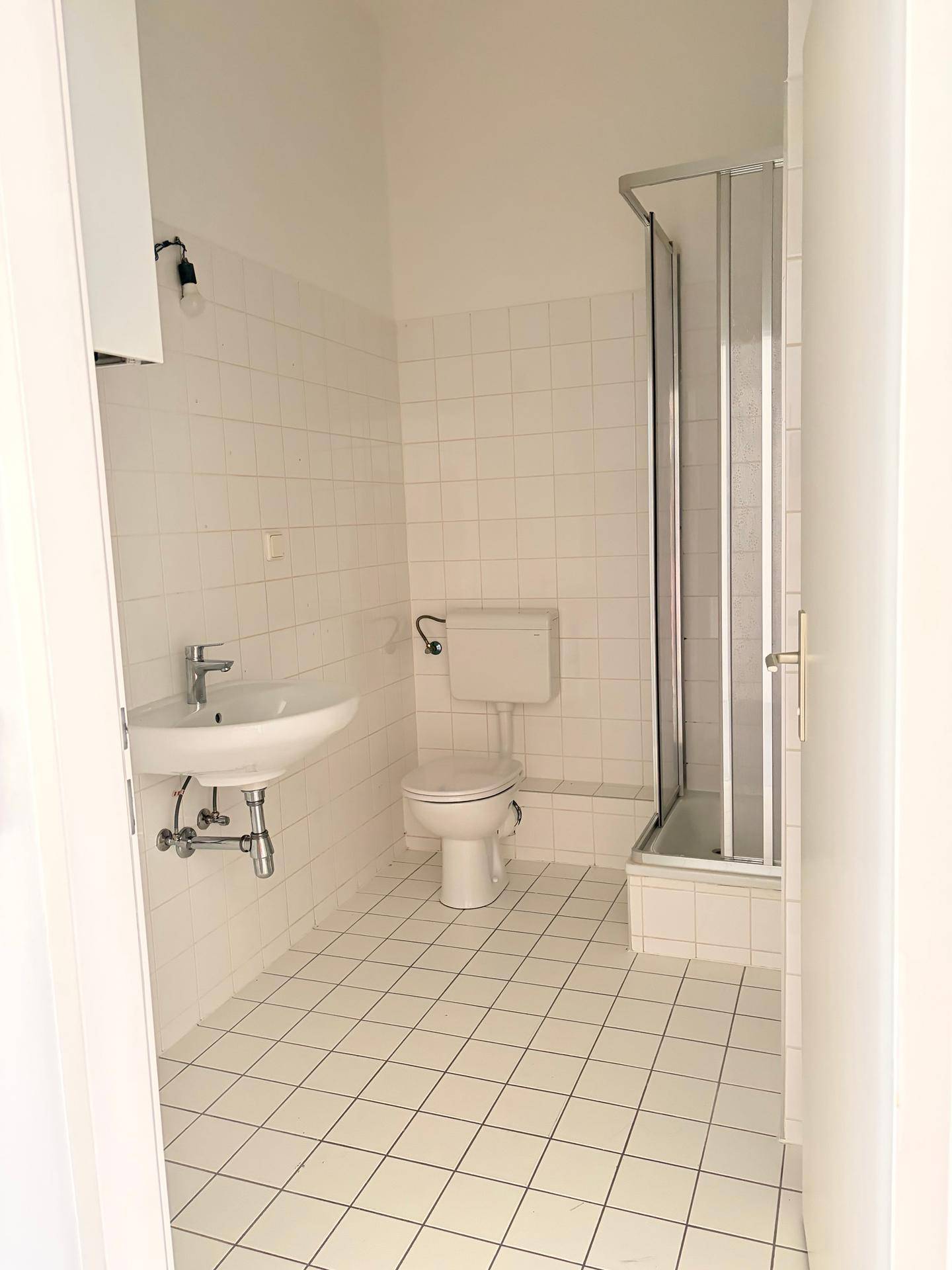 Helles Badezimmer mit weißen Fliesen, Waschbecken, Toilette und einer praktischen Duschkabine.