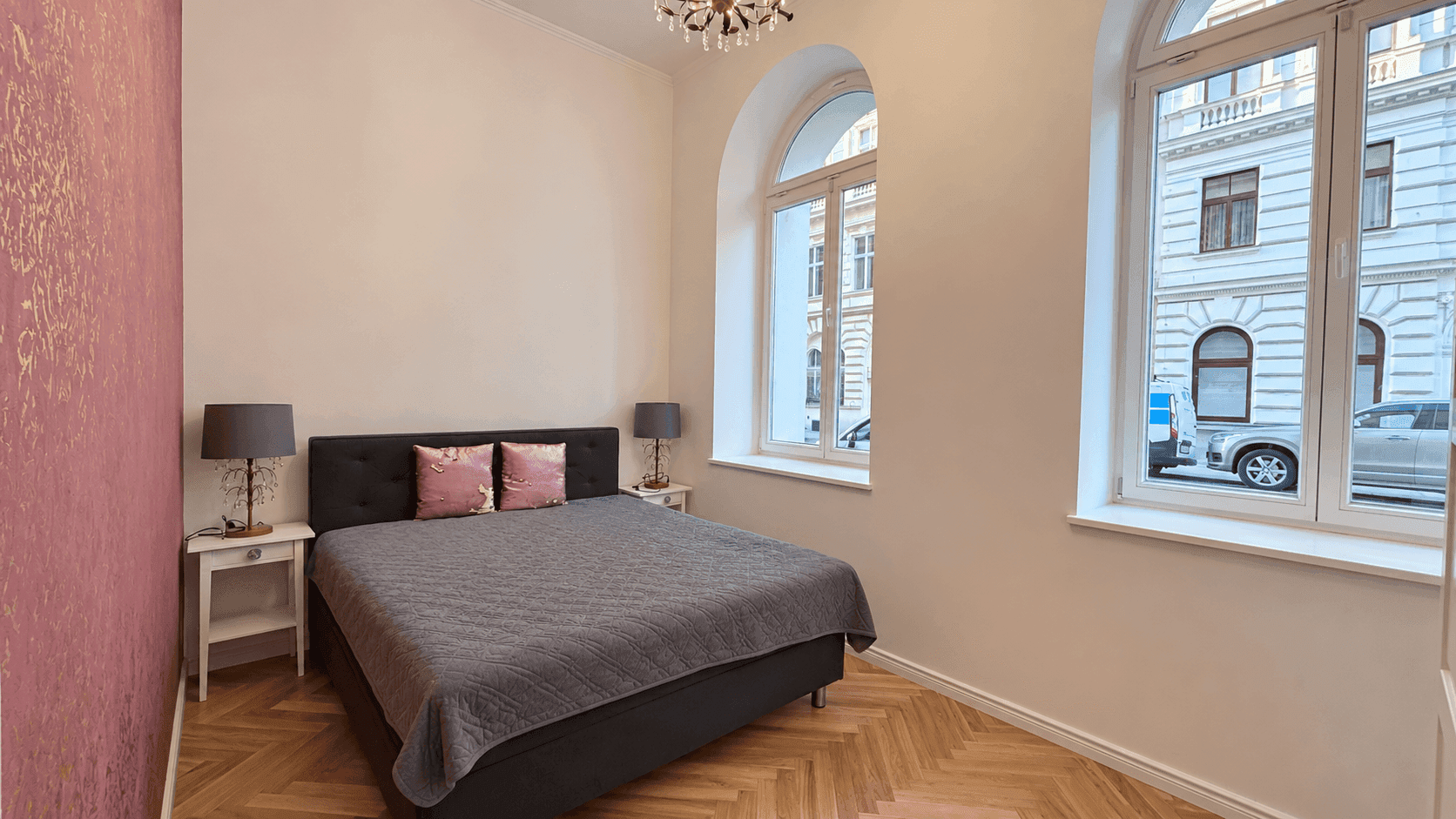 Helles Schlafzimmer mit Doppelbett, Nachttischen und zwei großen Rundbogenfenstern mit Blick auf die Straße.
