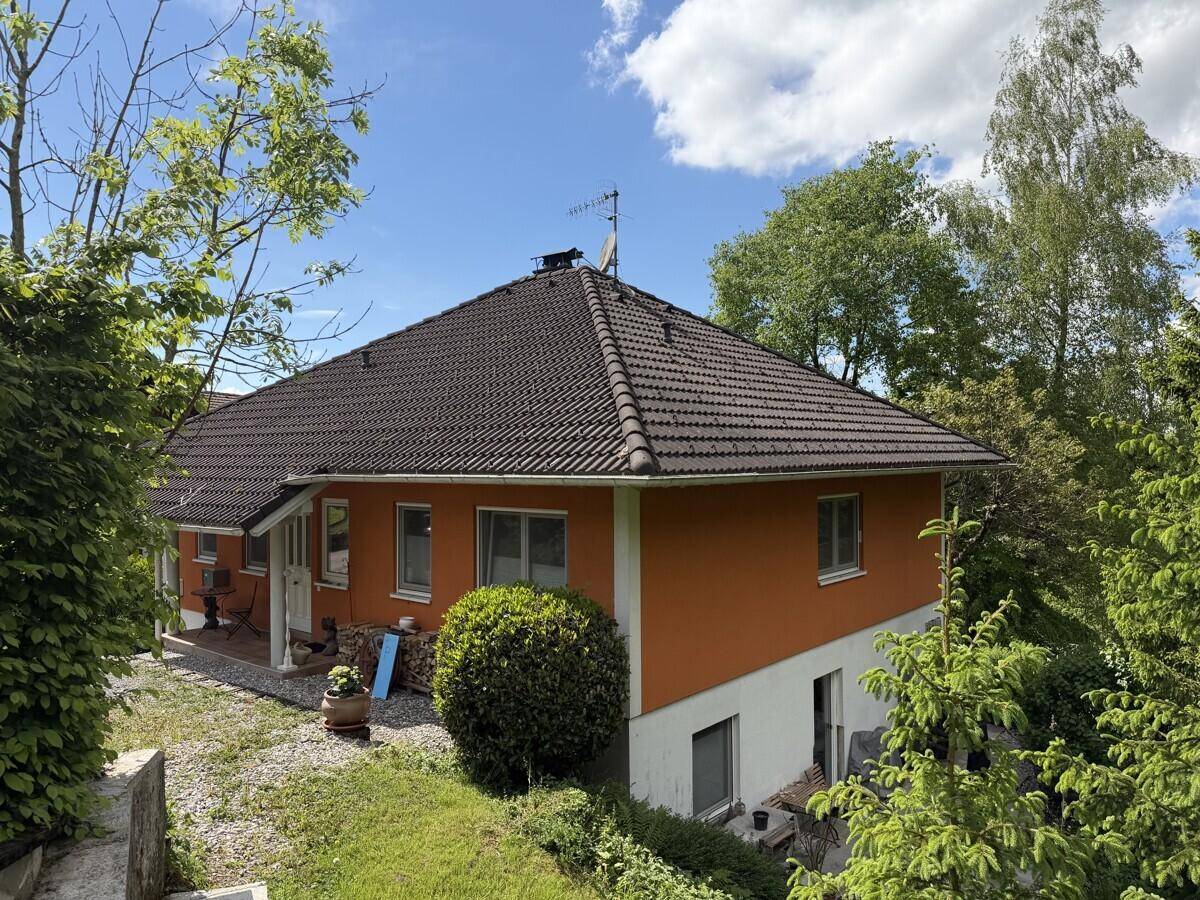 Wohnhaus mit orangefarbener Fassade, braunem Ziegeldach und Fenstern, umgeben von viel Grün.