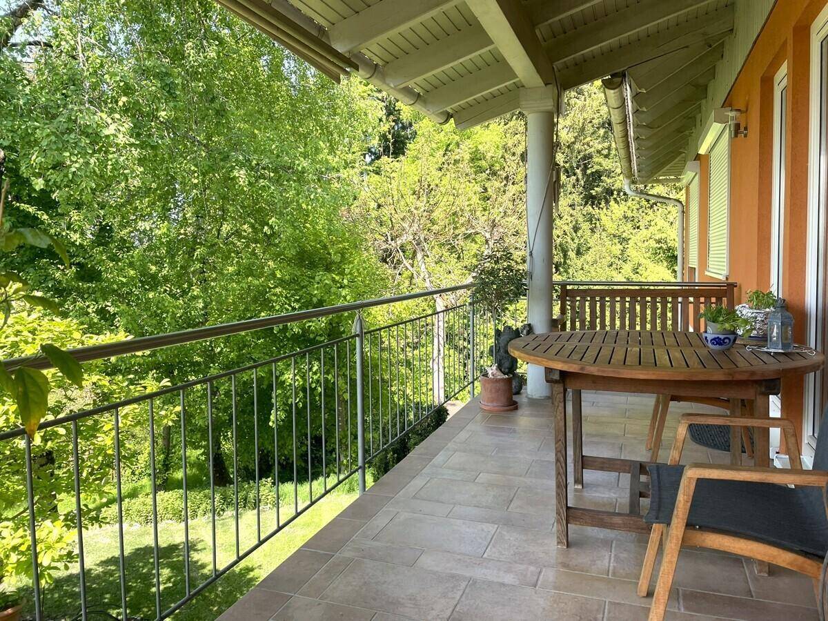 Geräumiger Balkon mit Fliesenboden, Metallgeländer und Holztisch, umgeben von üppigem Grün.