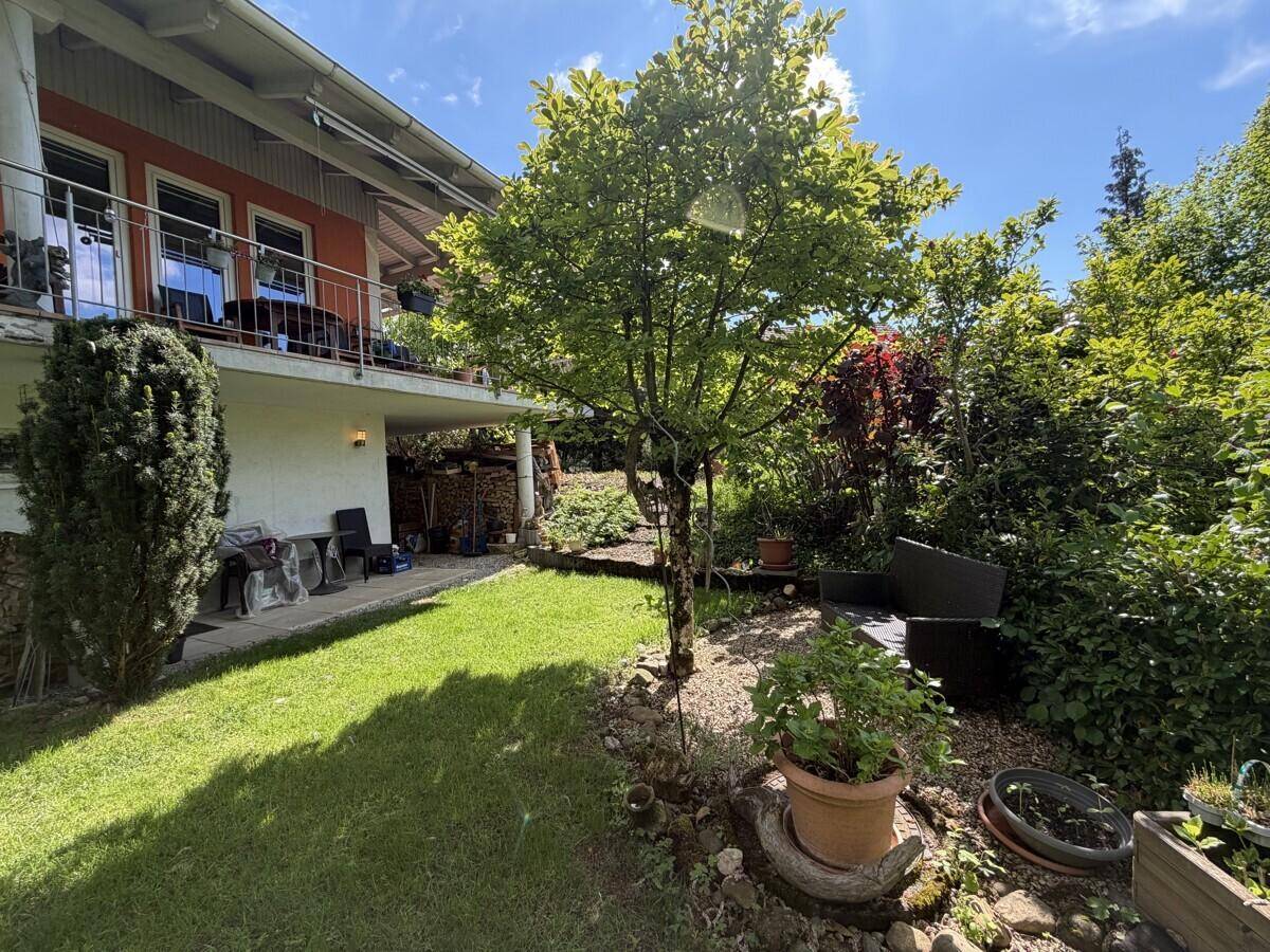 Heller Gartenbereich mit Rasen, einem Baum und einer kleinen Terrasse unter dem Balkon.