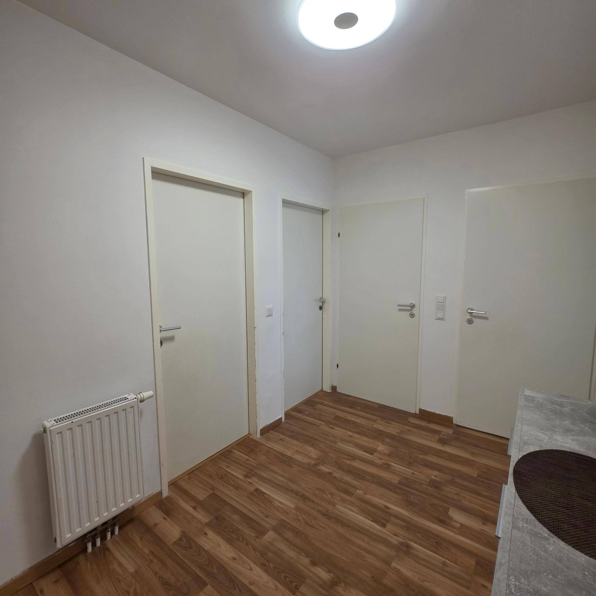 Schlichter Flur mit Holzfußboden, weiß gestrichenen Wänden und Heizkörper.