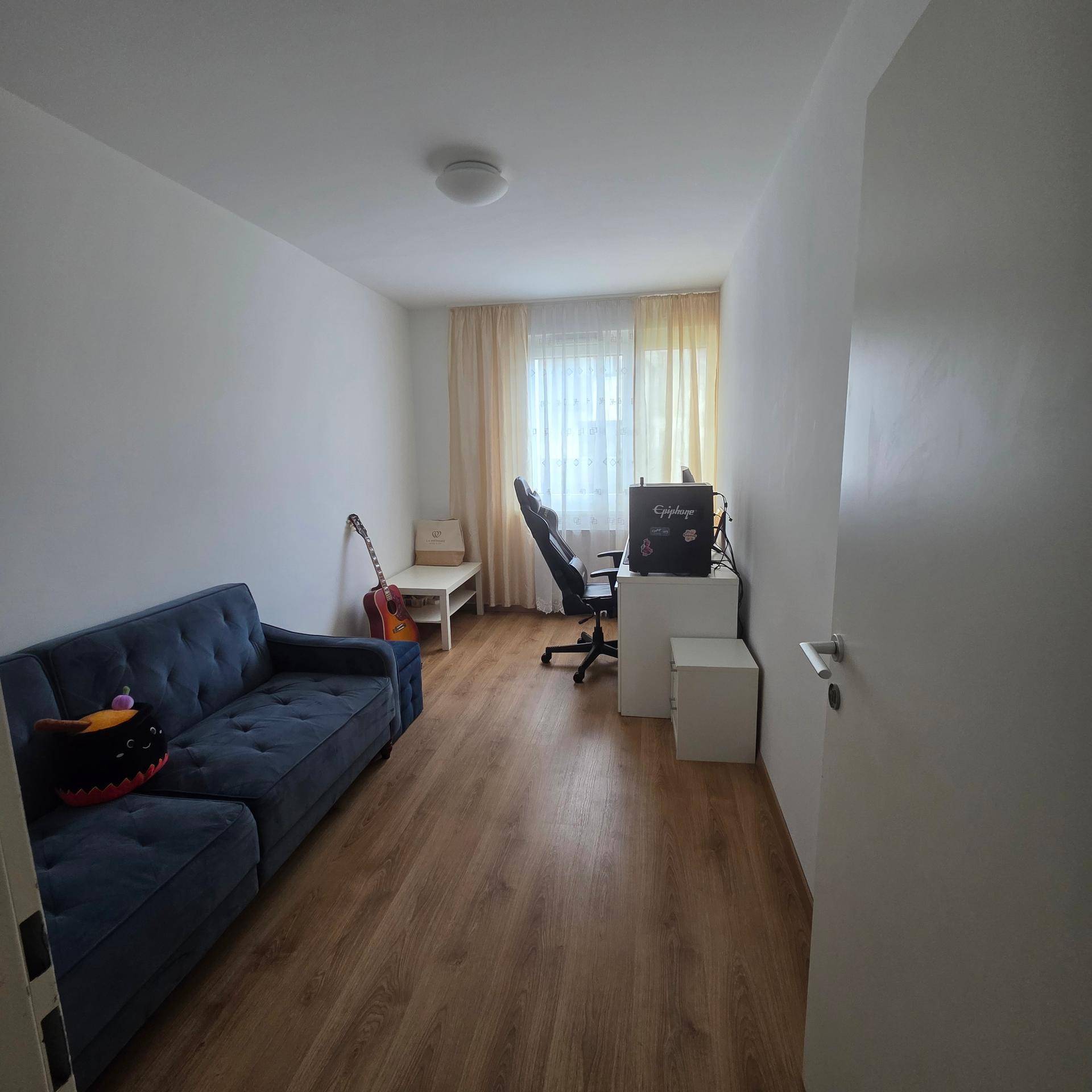 Helles Zimmer mit Holzfußboden, Sofa, Schreibtisch und Fenster mit Vorhängen.