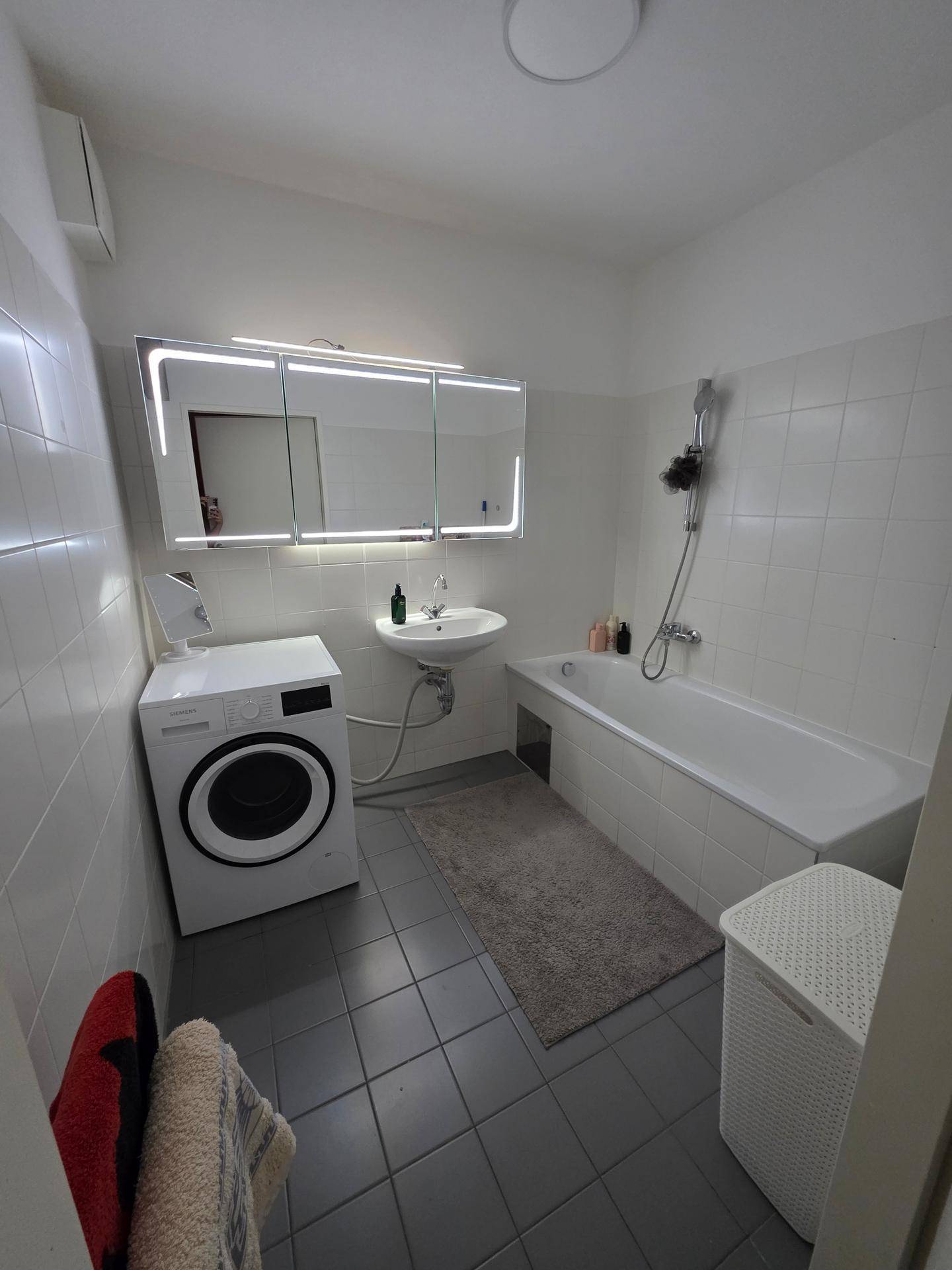 Modernes Badezimmer mit Badewanne, Waschbecken, Waschmaschine und beleuchtetem Spiegelschrank.