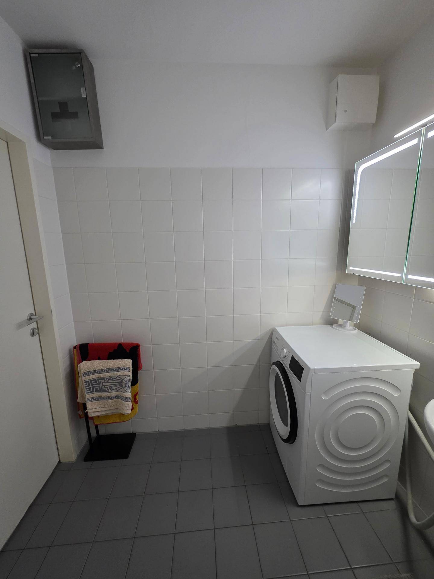 Badezimmer mit Waschmaschine, Fliesen und beleuchtetem Spiegelschrank.
