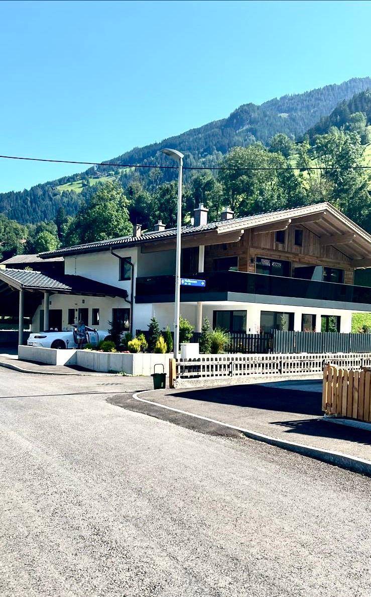 Modernes Haus mit Holzfassade, Balkonen und Carport, eingebettet in eine grüne Berglandschaft.