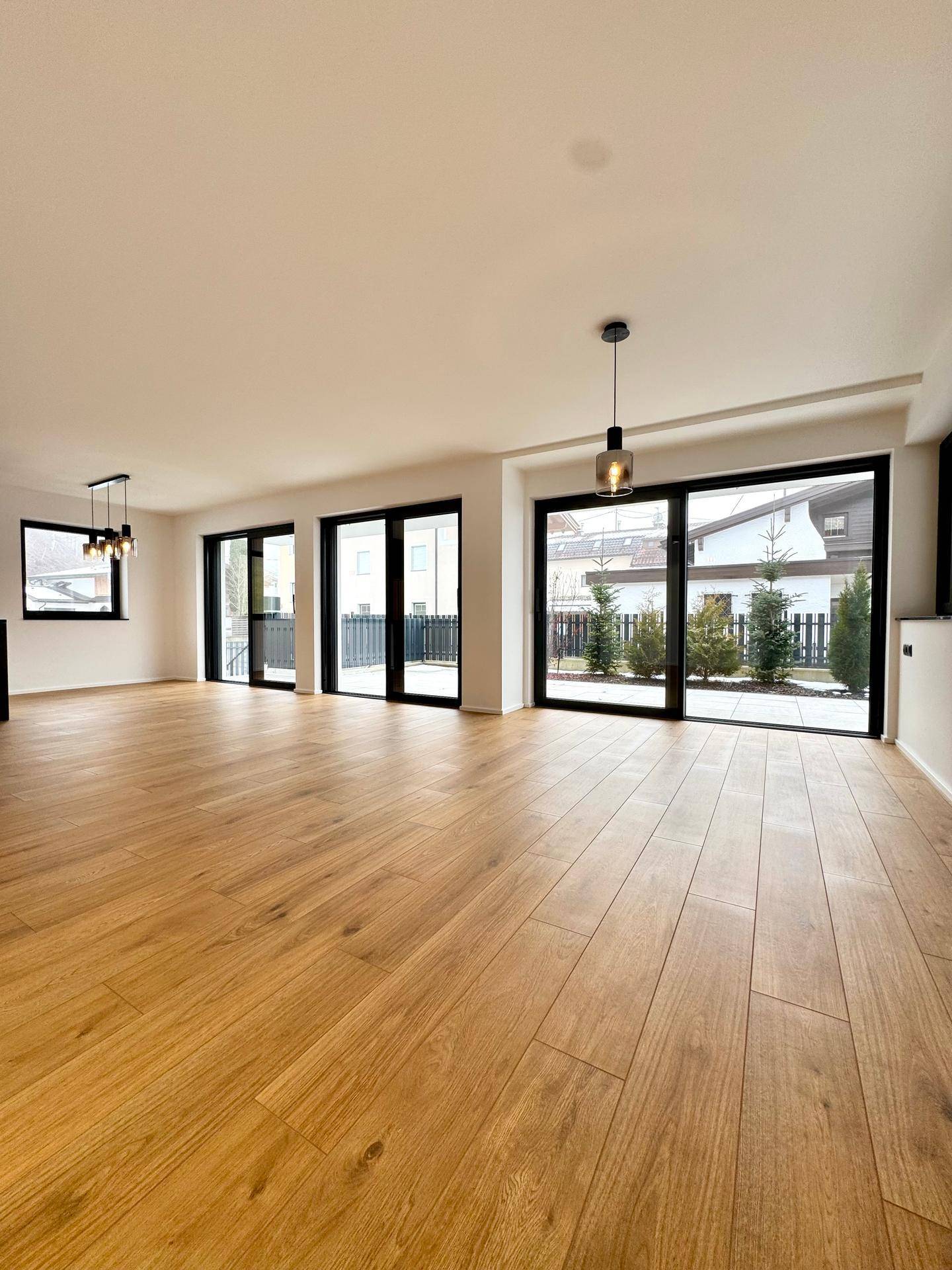 Heller, leerer Wohnbereich mit modernem Holzboden und großen Fenstern mit Zugang zur Terrasse.