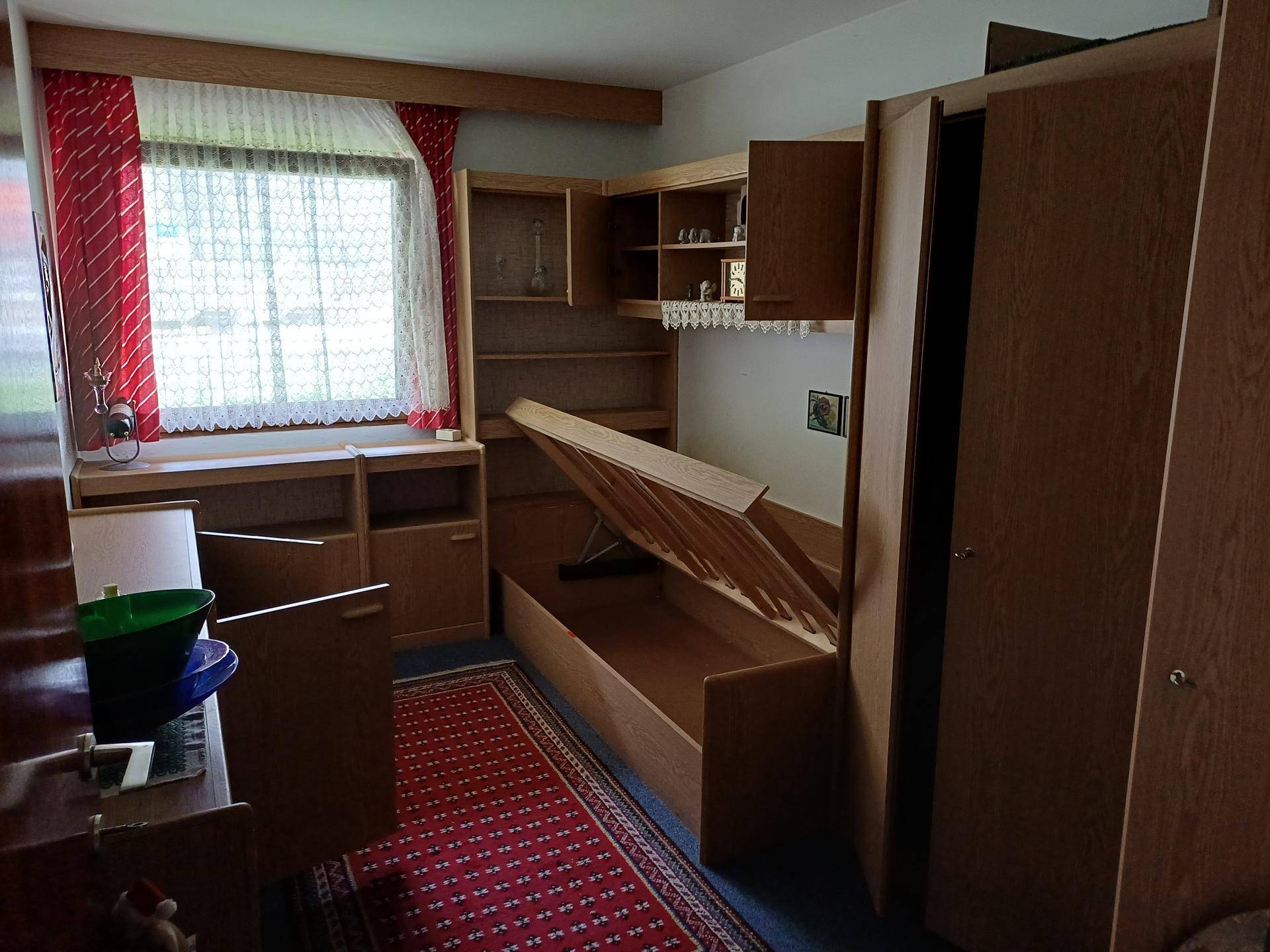 Schlafzimmer mit Einzelbett, Holzmöbeln und einem Fenster mit rotem Vorhang.