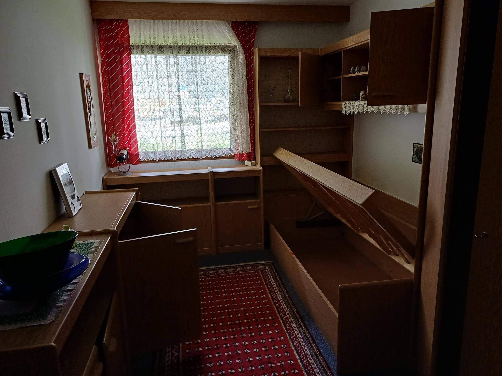 Schlafzimmer mit Einzelbett, Holzmöbeln und einem Fenster mit rotem Vorhang.