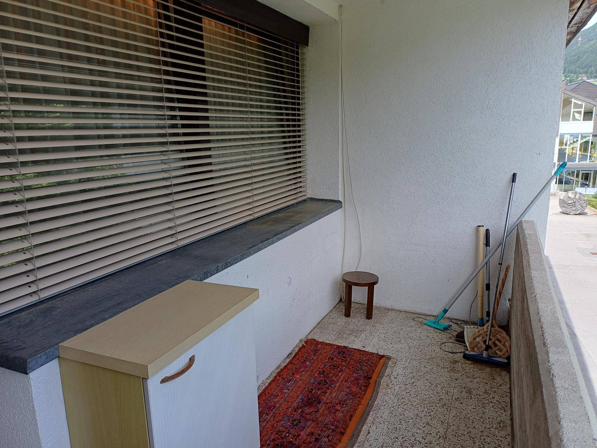 Schmaler Balkon mit Jalousien, einem kleinen Hocker und Reinigungsgeräten.