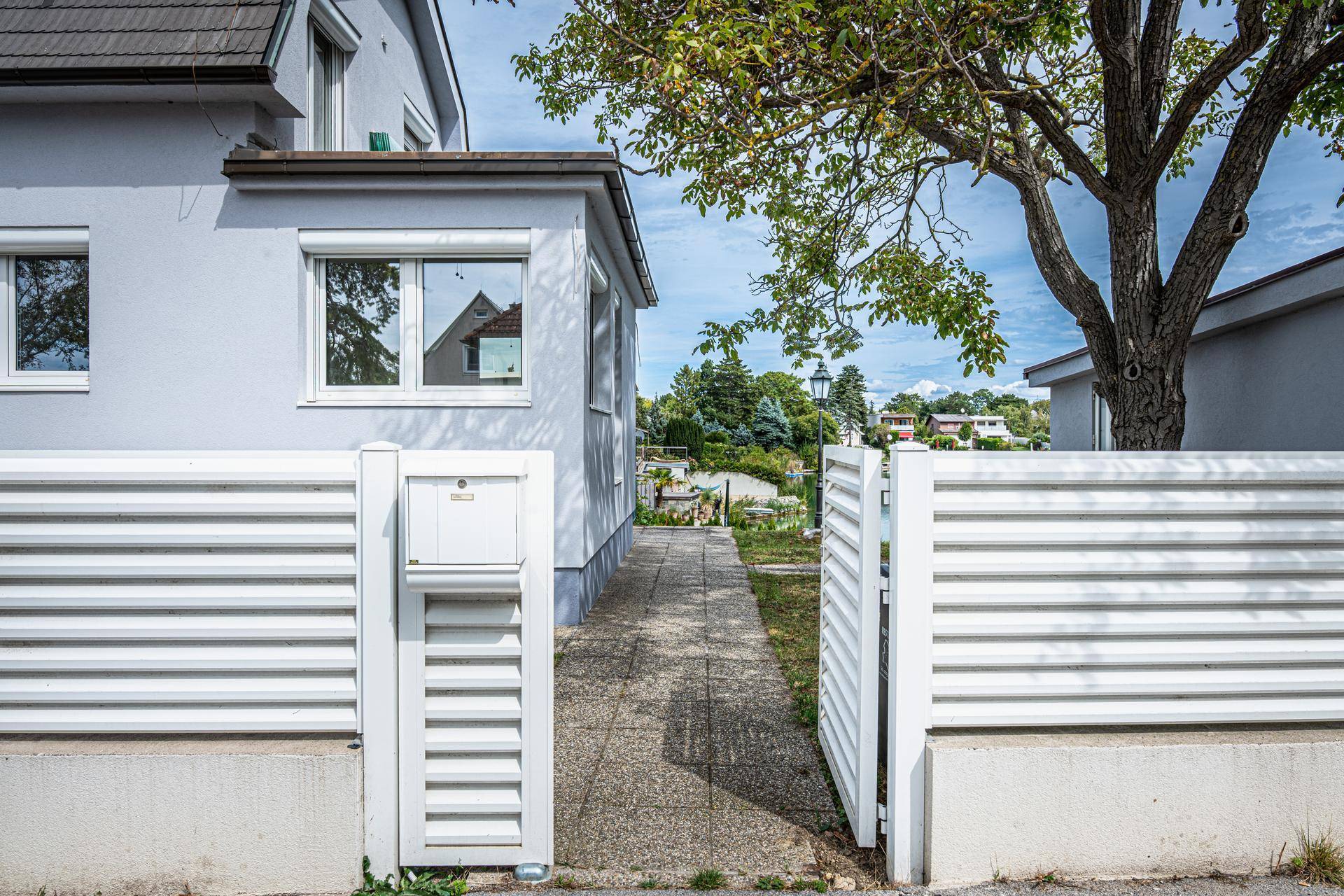 Eingangsbereich des Hauses mit weißem Zaun und Briefkasten, umgeben von Grün.