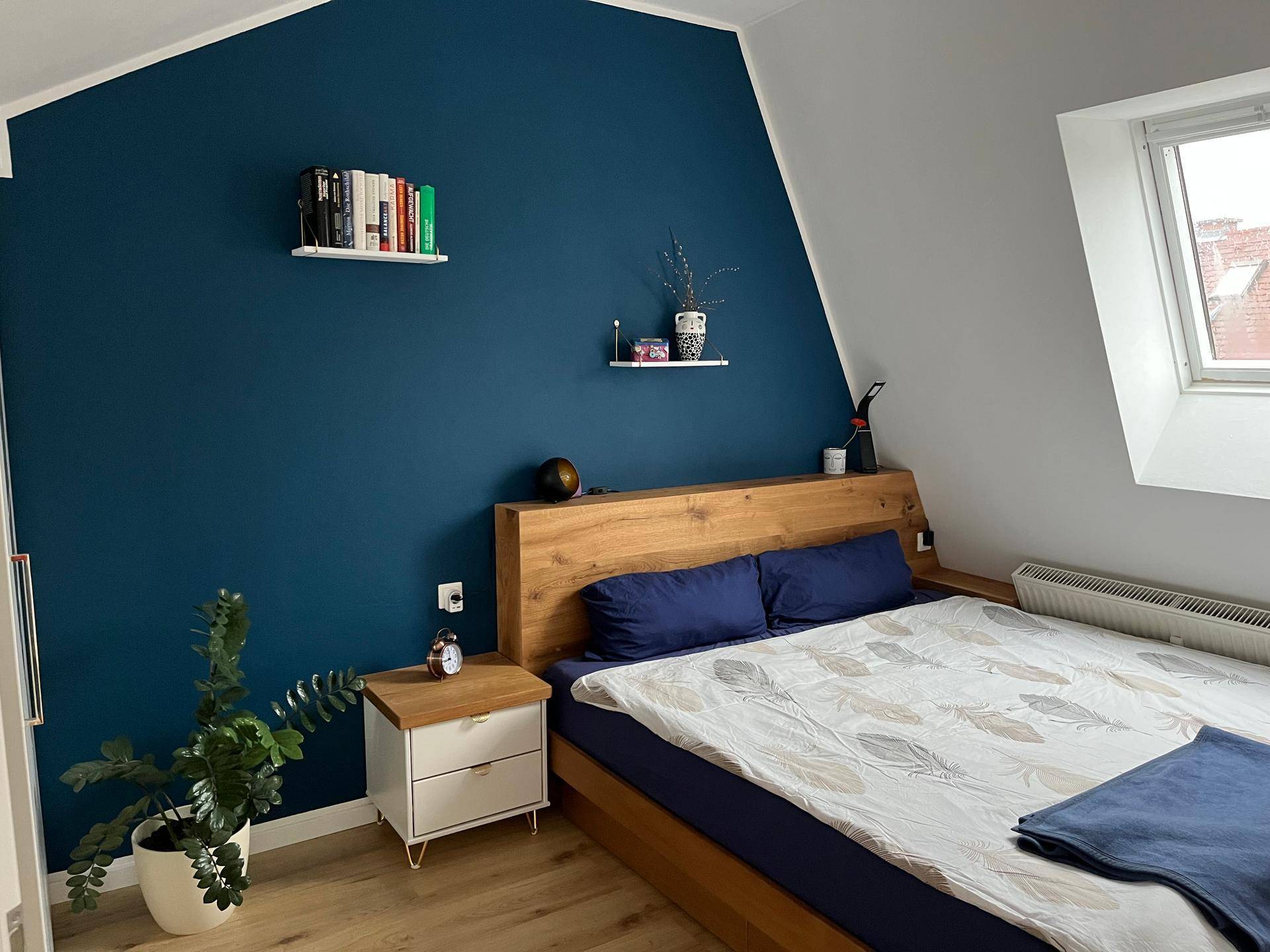 Gemütliches Schlafzimmer mit dunkelblauer Akzentwand, Holzbett und Dachfenster.