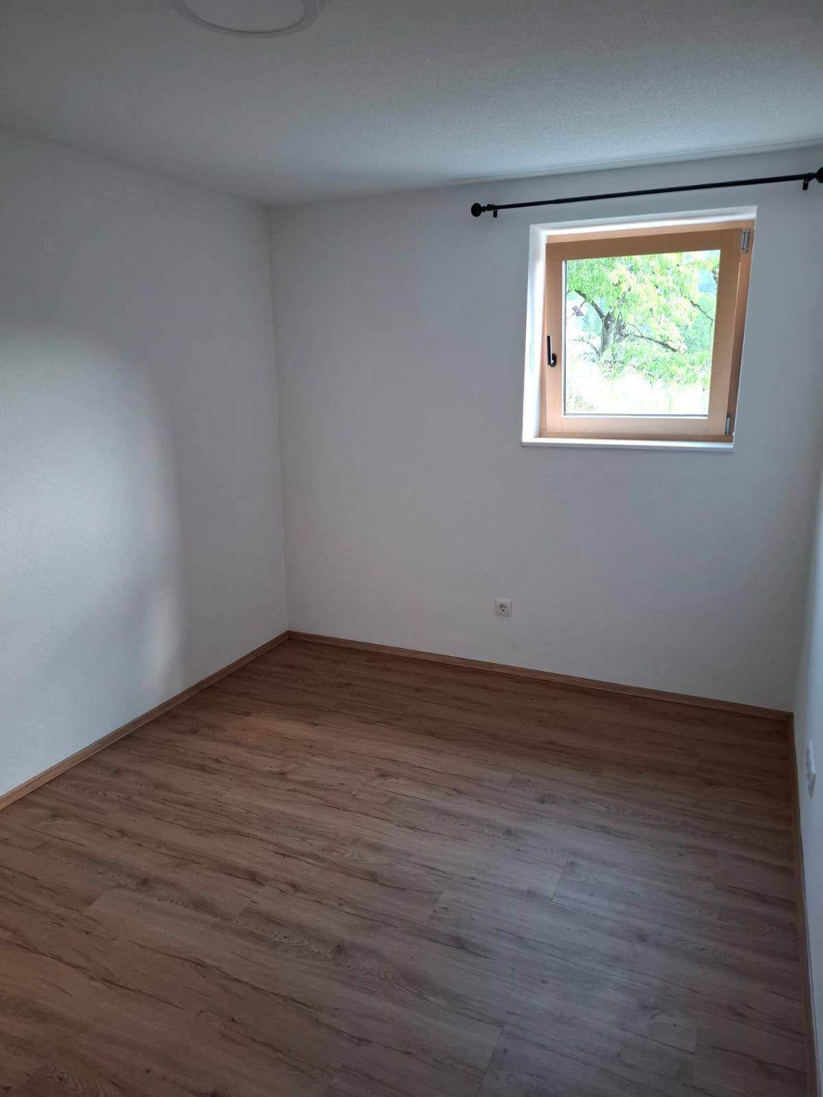 Helles, leeres Zimmer mit Laminatboden und einem Fenster mit Blick ins Grüne.