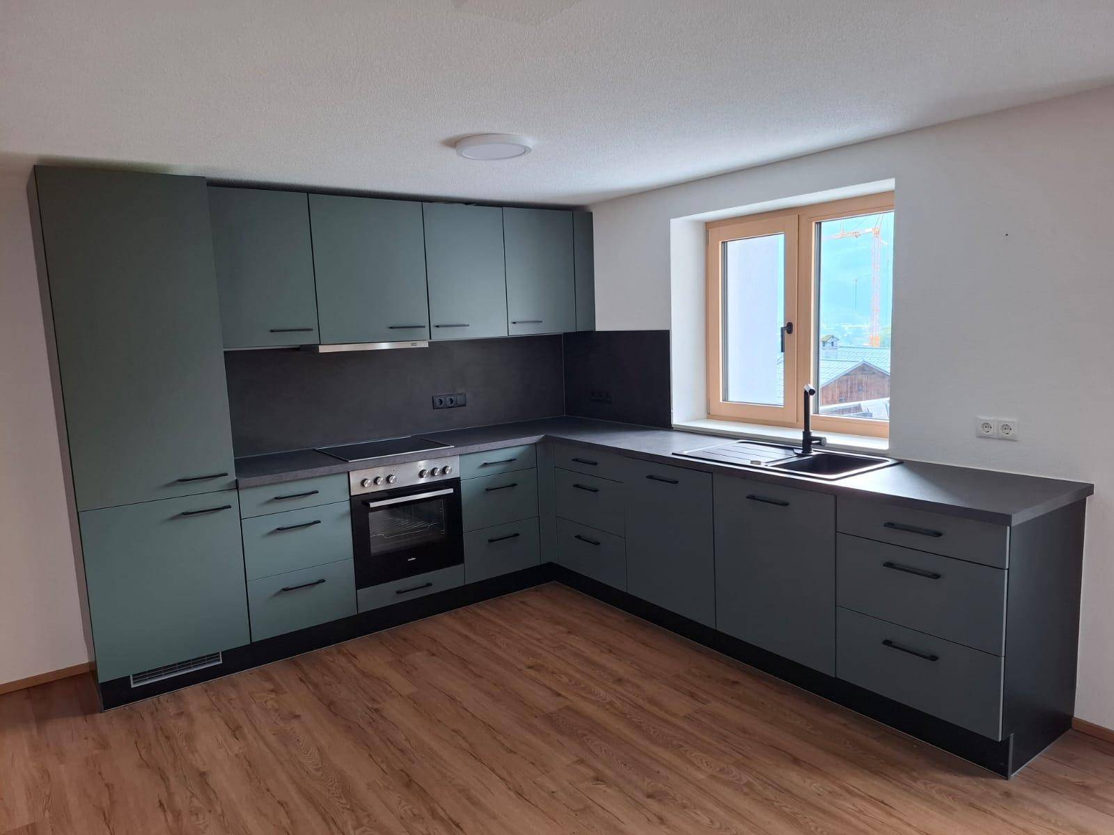 Moderne Küche mit dunkelgrünen Schränken, Einbauherd und Spüle unter dem Fenster.