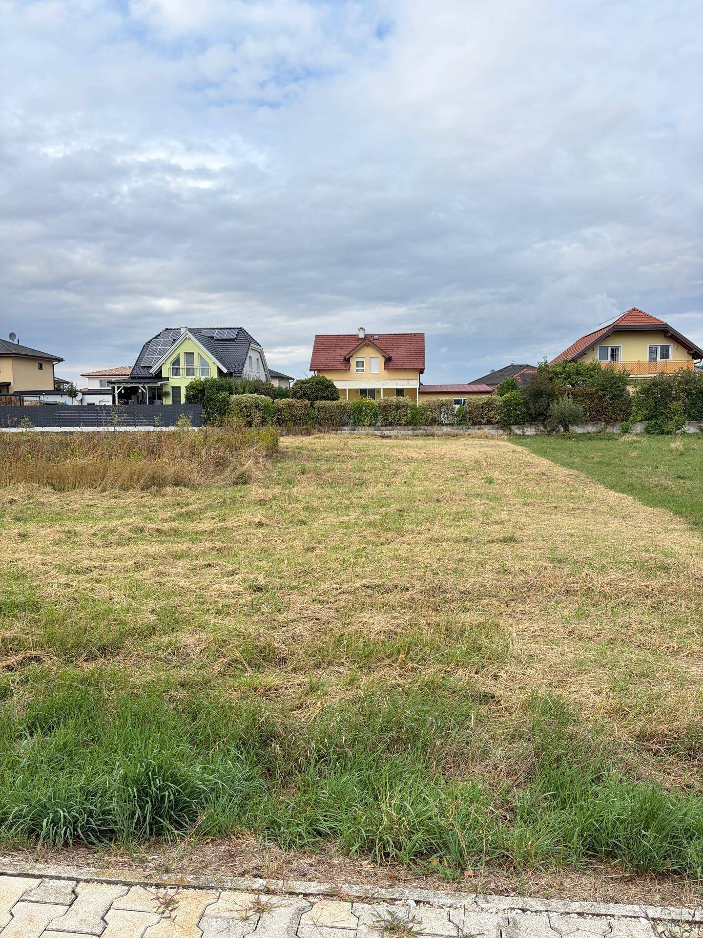 Leeres Grundstück mit Grasfläche und Wohnhäusern im Hintergrund unter bewölktem Himmel.