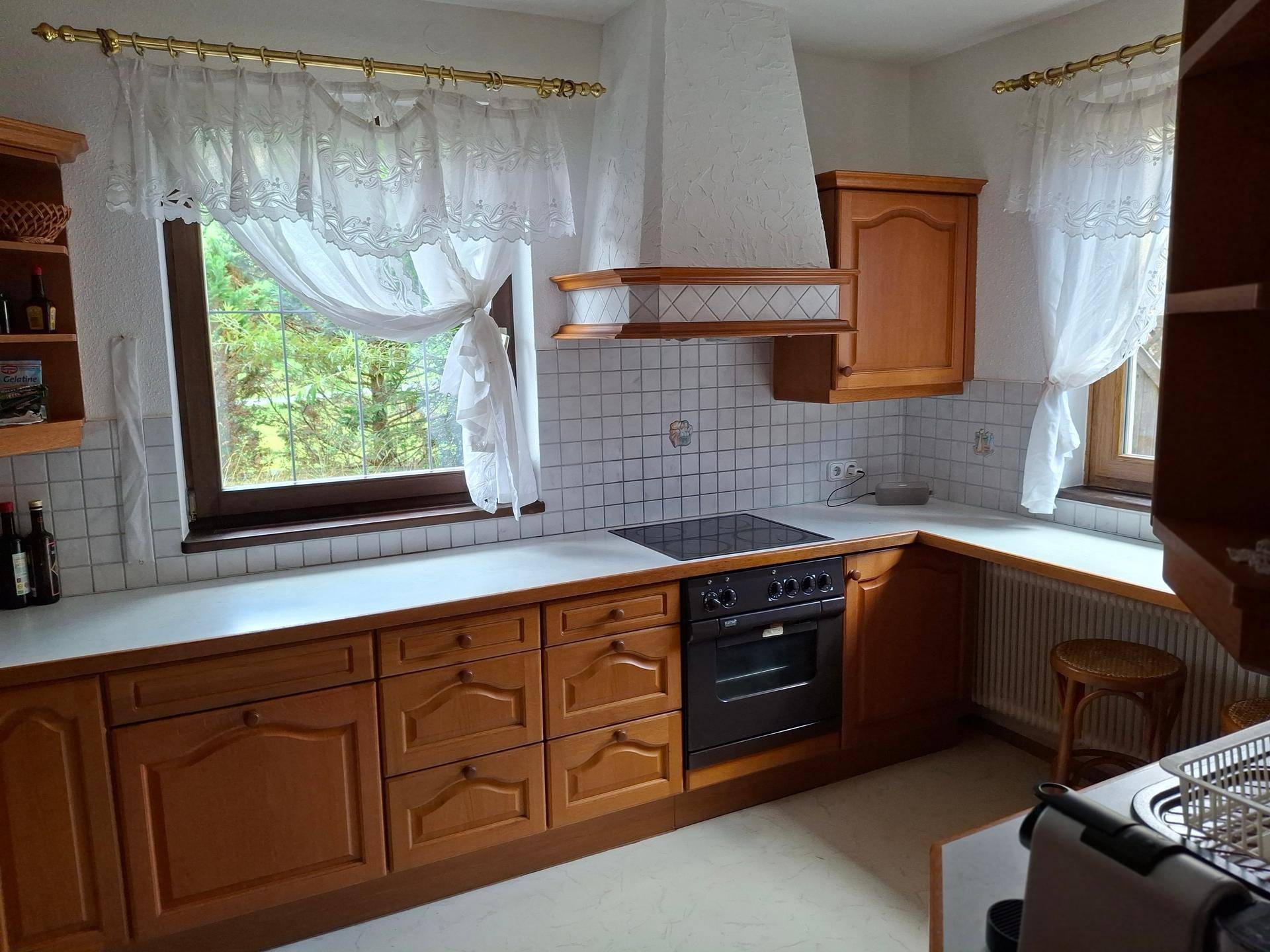 Traditionelle Holzküche mit weißen Arbeitsplatten, Kochfeld und Fenstern mit Spitzenvorhängen.