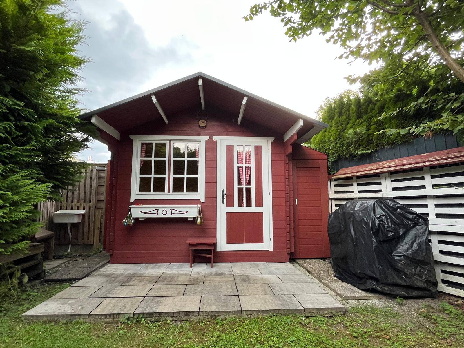 Roter Holzgartenpavillon mit kleiner Terrasse, umgeben von Hecken und einem Außenwaschbecken.