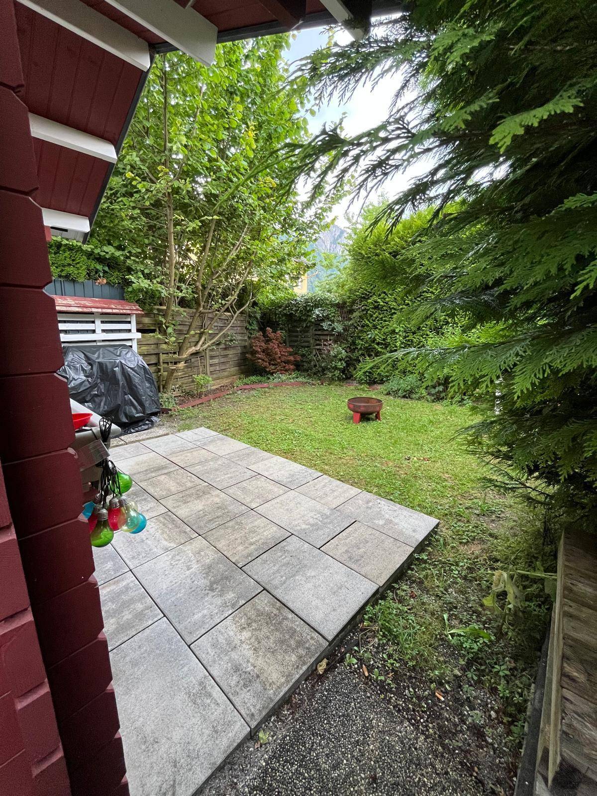 Blick von der überdachten Terrasse auf den Garten mit Rasenfläche und Feuerstelle im Hintergrund.