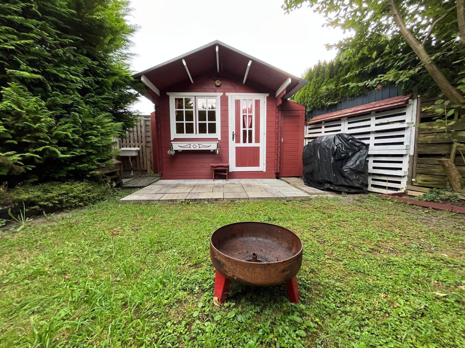 Gartenbereich mit rotem Holzpavillon, gepflasterter Terrasse und einer Feuerstelle auf dem Rasen.