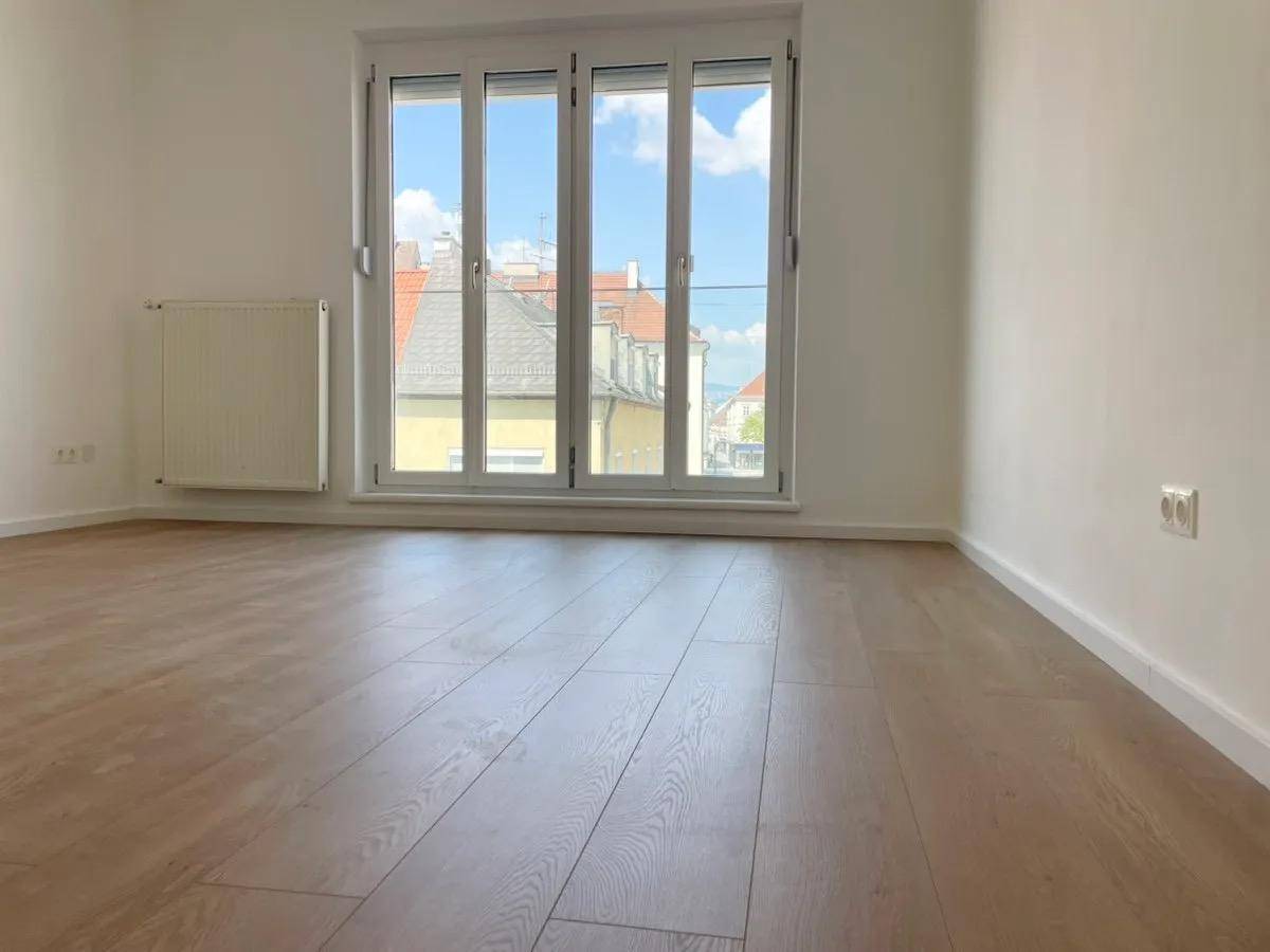 Geräumiges Zimmer mit Holzboden und großem Fenster, das einen weiten Blick über die Dächer bietet.