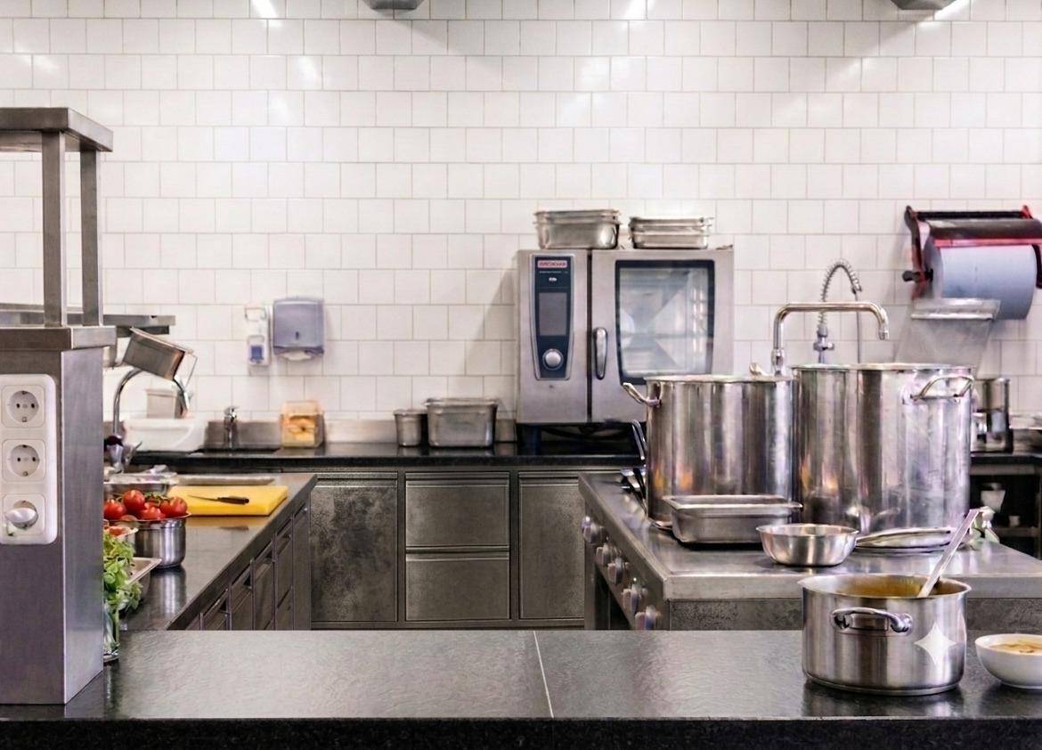 Detailansicht einer professionellen Küche mit großen Kochtöpfen, Edelstahlflächen und weißen Fliesen.