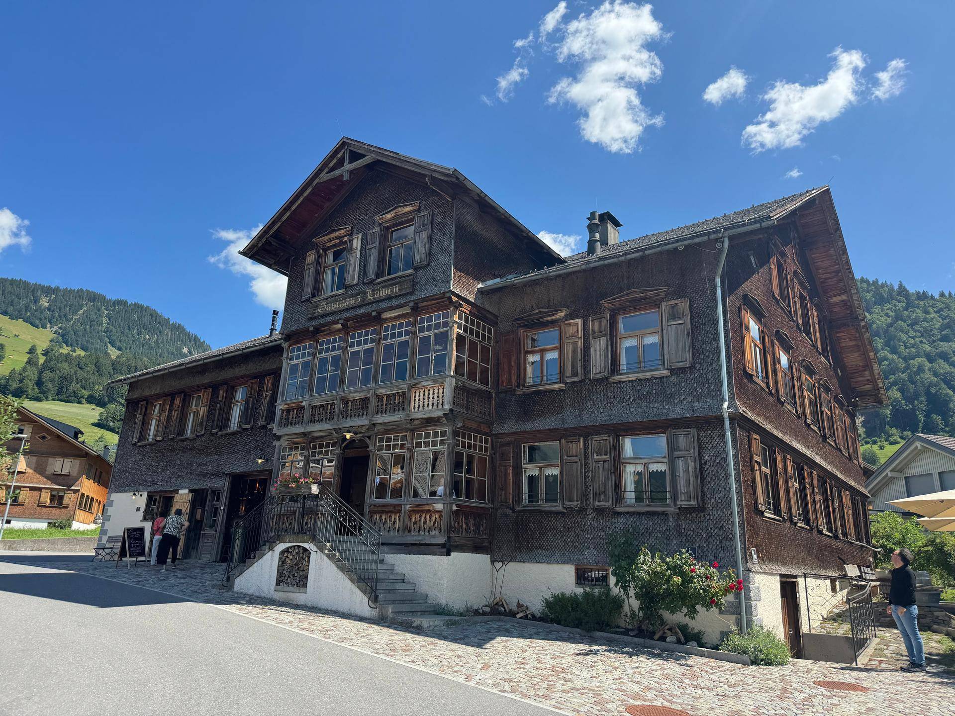 Traditionelles Holzgebäude mit vielen Fenstern und einer komplexen Dachstruktur vor einer Bergkulisse.