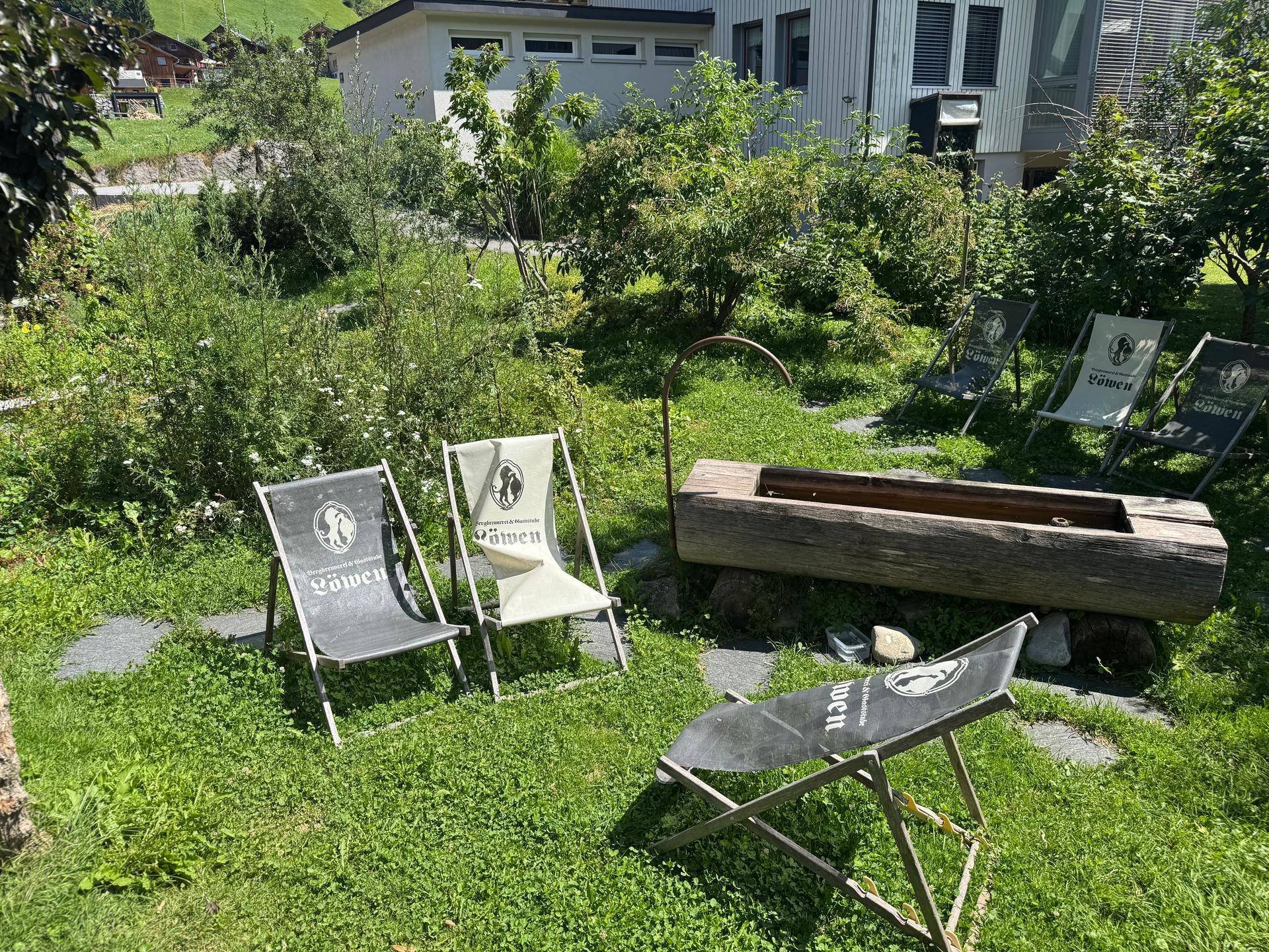 Gartenbereich mit Liegestühlen und einer hölzernen Feuerstelle, umgeben von üppigem Grün.