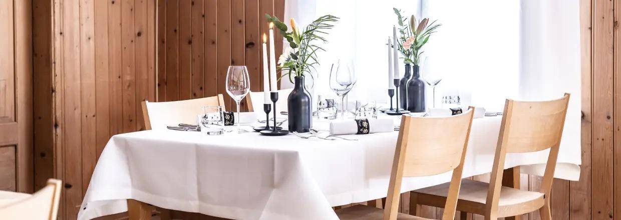 Elegant gedeckter Tisch mit weißer Tischdecke, Kerzen und Blumenarrangements in einem Holzraum.
