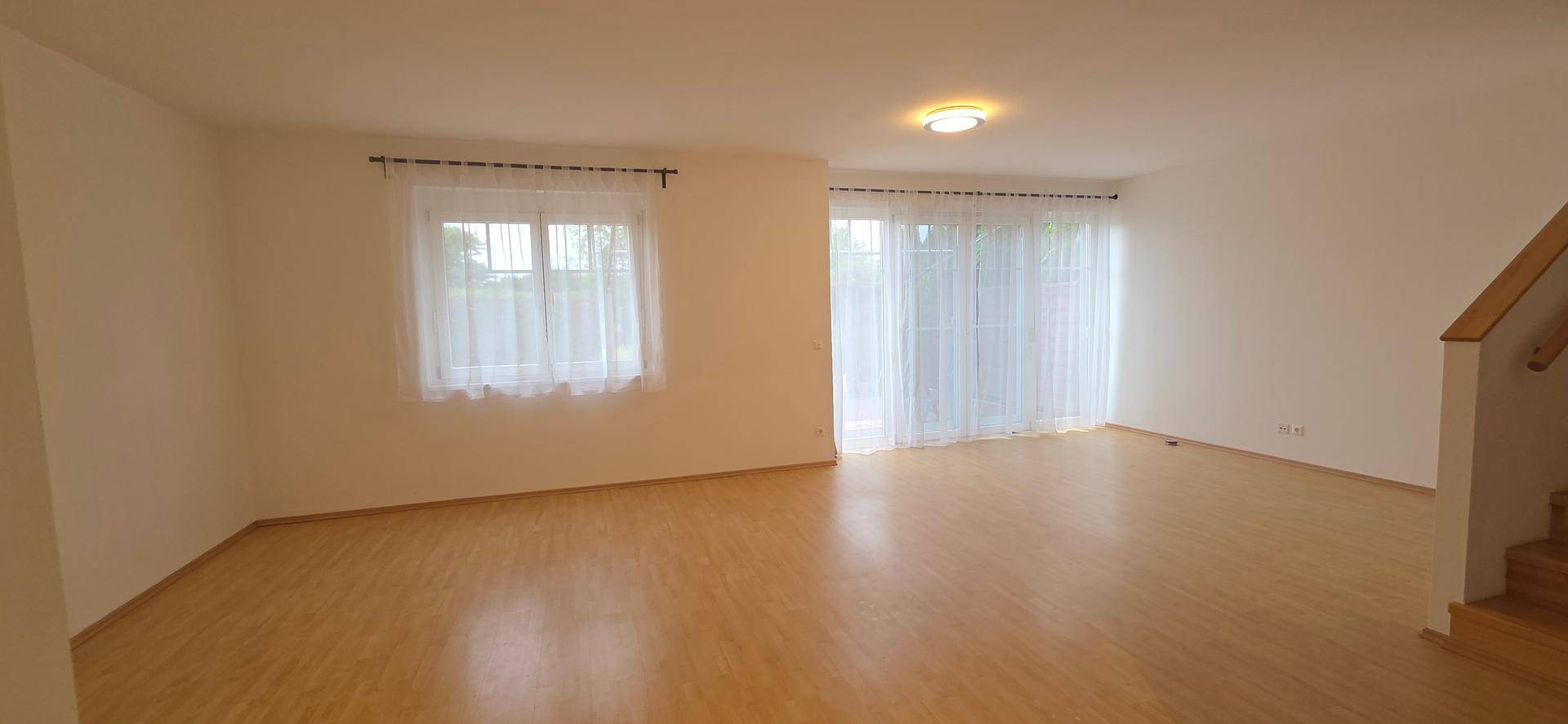 Geräumiges Wohnzimmer mit hellem Laminatboden und großen Fenstern, die viel Licht hereinlassen.