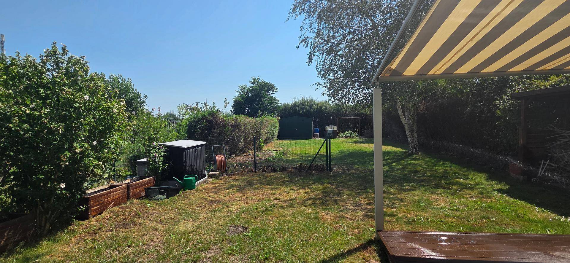 Gepflegter Gartenbereich mit Rasenfläche, Sträuchern und einer gestreiften Markise für Schatten.