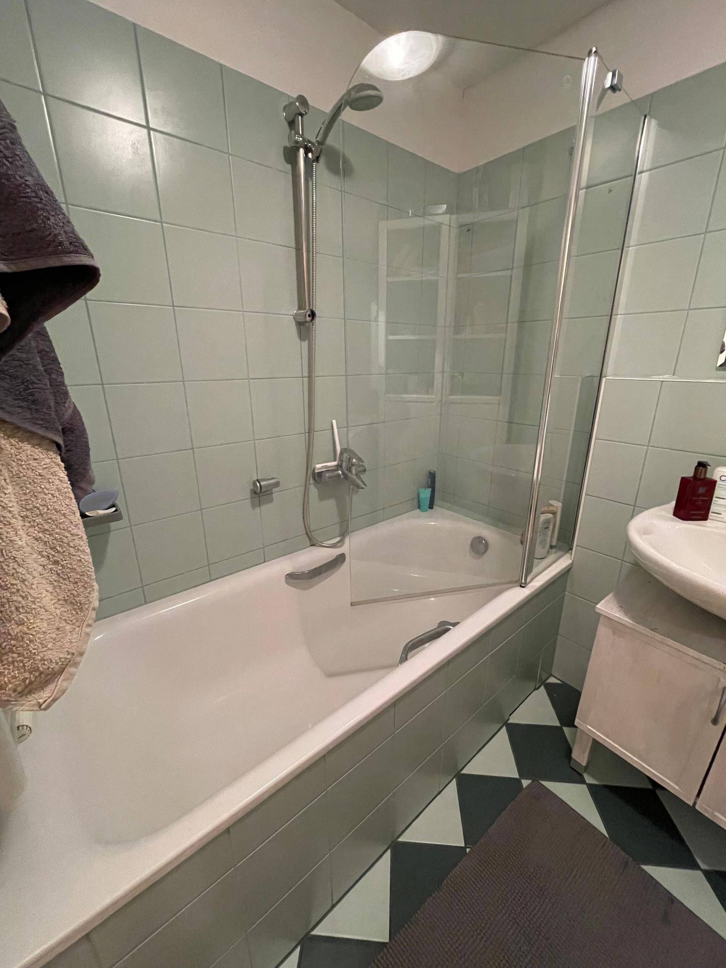 Badezimmer mit Badewanne und Duschabtrennung, hellgrünen Fliesen und kariertem Boden, funktional gestaltet.