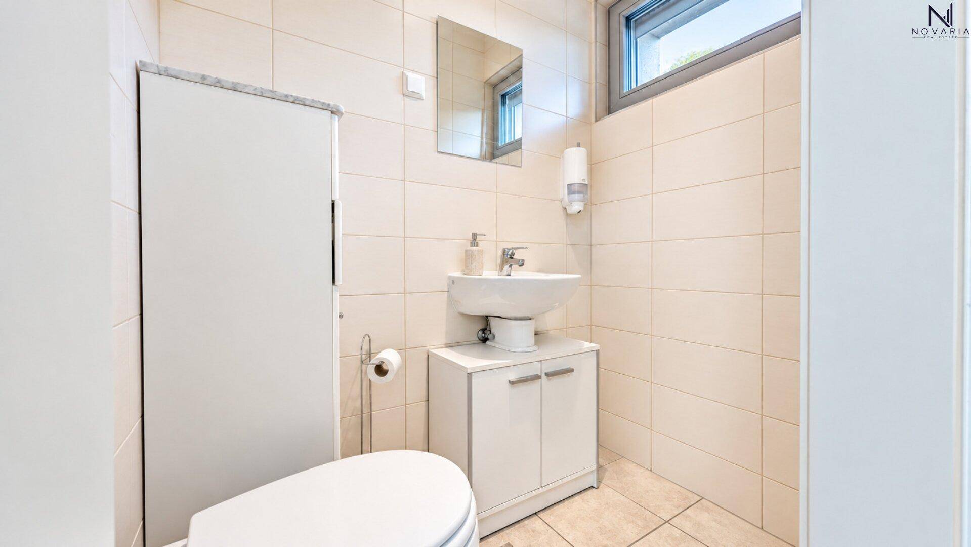 Kleines Gäste-WC mit Fenster, Waschbecken und wandmontierter Toilette in hellen Fliesen.