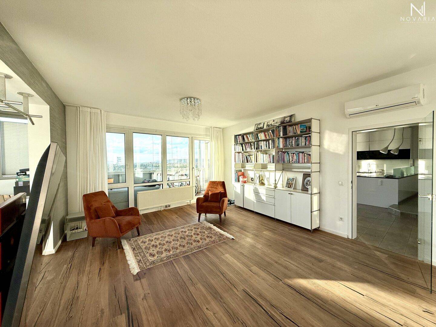 Helles Wohnzimmer mit Holzboden, Bücherregal und Zugang zu einer modernen Küche.