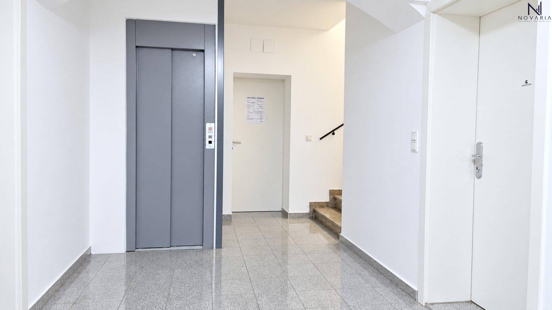Eingangsbereich mit Aufzug und Treppenhaus, ausgestattet mit Fliesenboden und weißen Wänden.