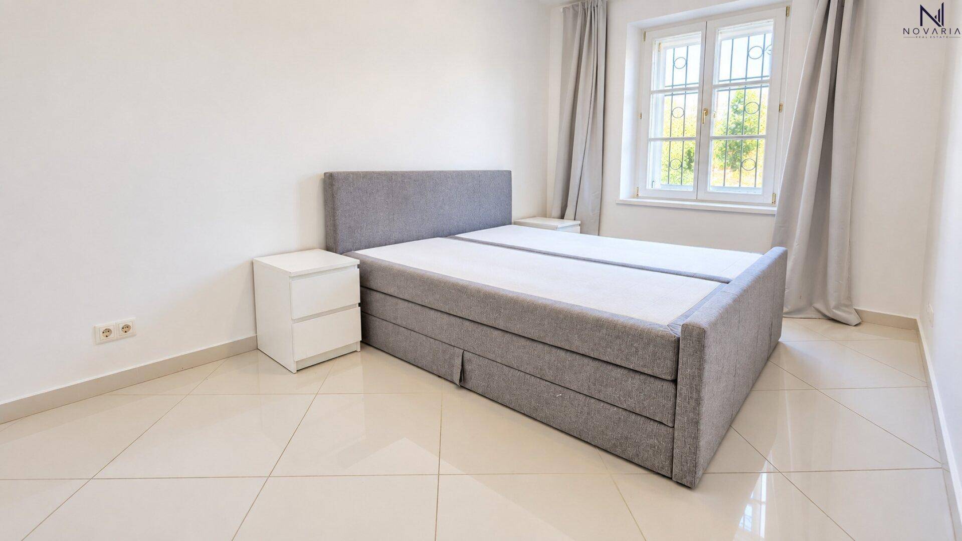 Schlafzimmer mit einem modernen grauen Boxspringbett, weißen Nachttischen und einem Fenster mit Vorhängen.