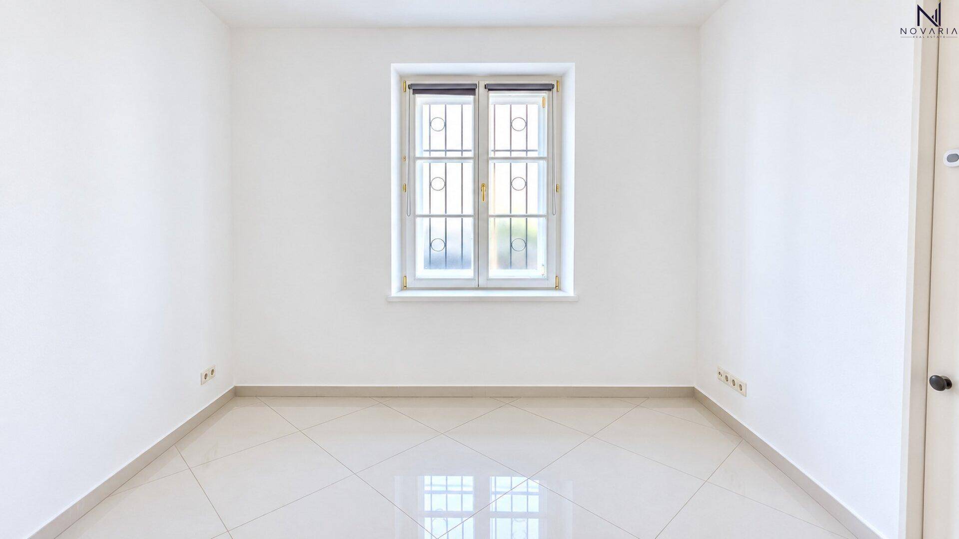 Leerer Raum mit weißen Wänden, Fliesenboden und einem Fenster mit Gitterstäben.