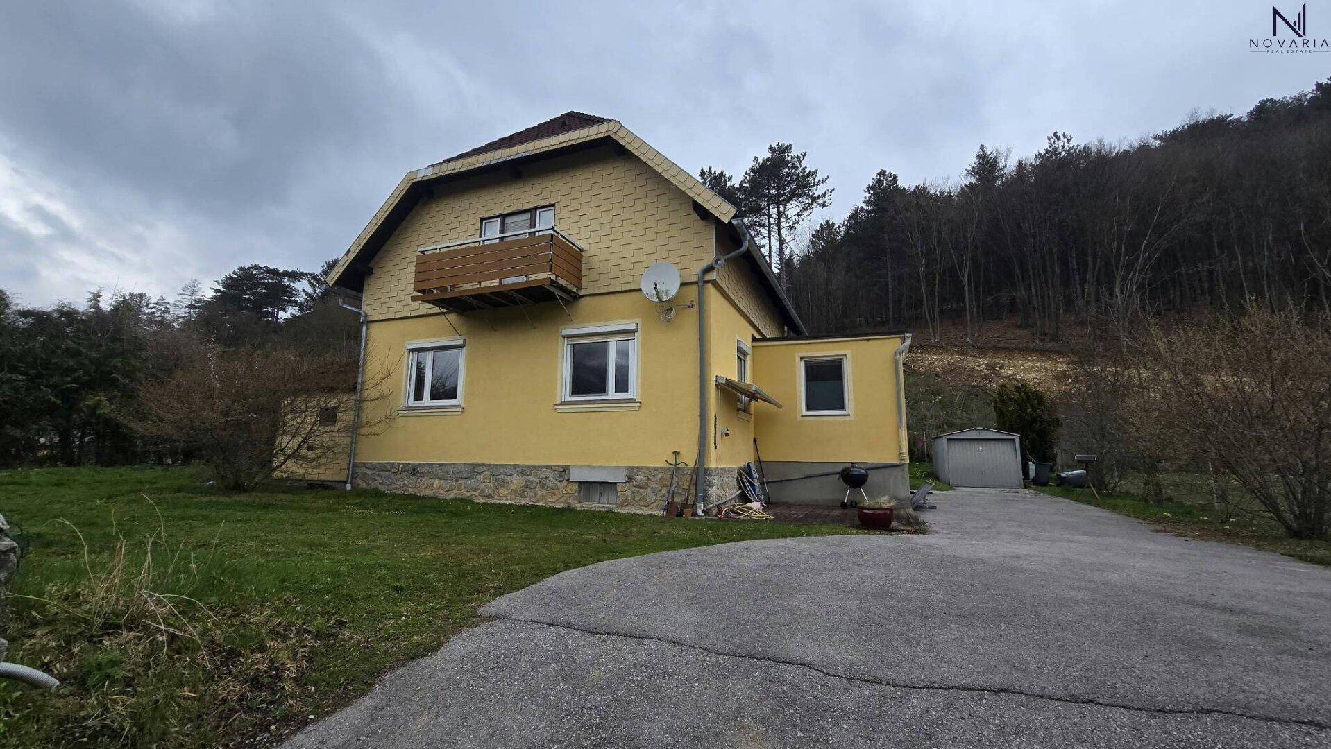 Gelbes Einfamilienhaus mit kleinem Holzbalkon und Zufahrt, umgeben von Bäumen unter bewölktem Himmel.