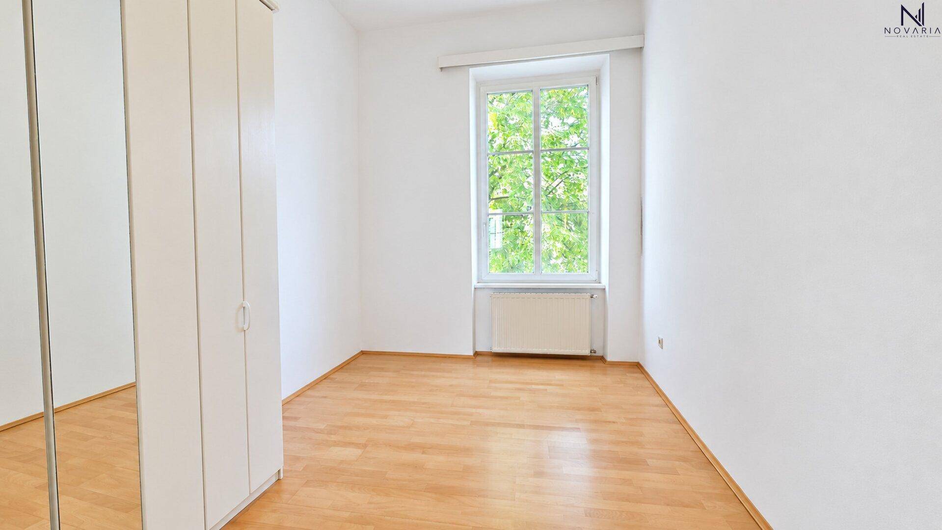 Schlafzimmer mit Parkettboden, weißem Kleiderschrank und Blick ins Grüne durch das Fenster.
