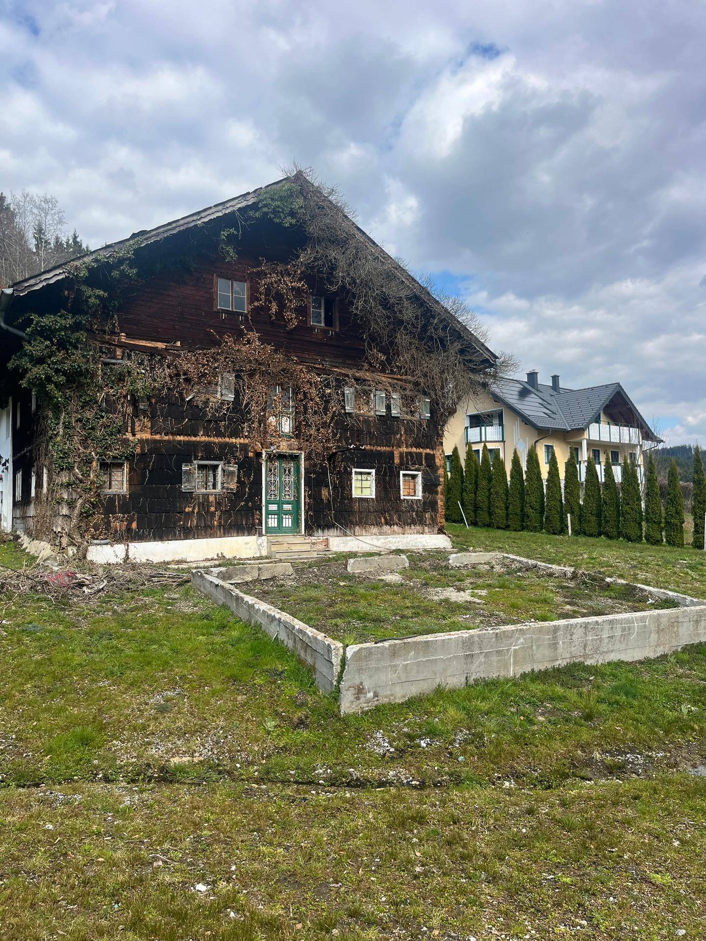 Vorderansicht des rustikalen Holzhauses mit grüner Tür und einer Betonumrandung im Vordergrund.