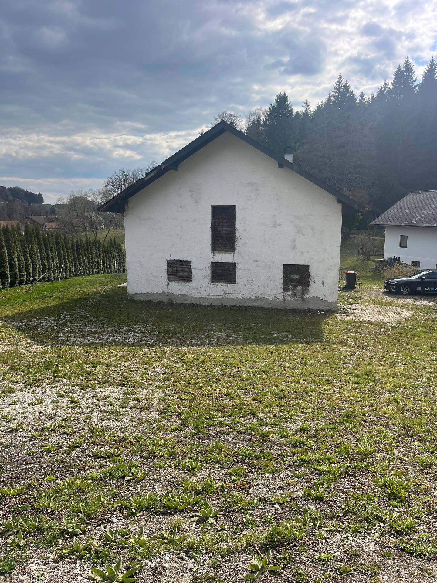 Kleines weißes Gebäude mit zugenagelten Fenstern, umgeben von einer Wiese und Bäumen.