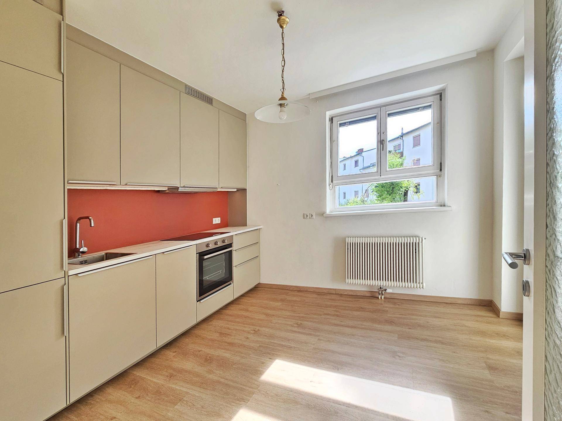 Helle Küche mit beigen Schränken, integriertem Backofen und Fenster mit Blick nach draußen.