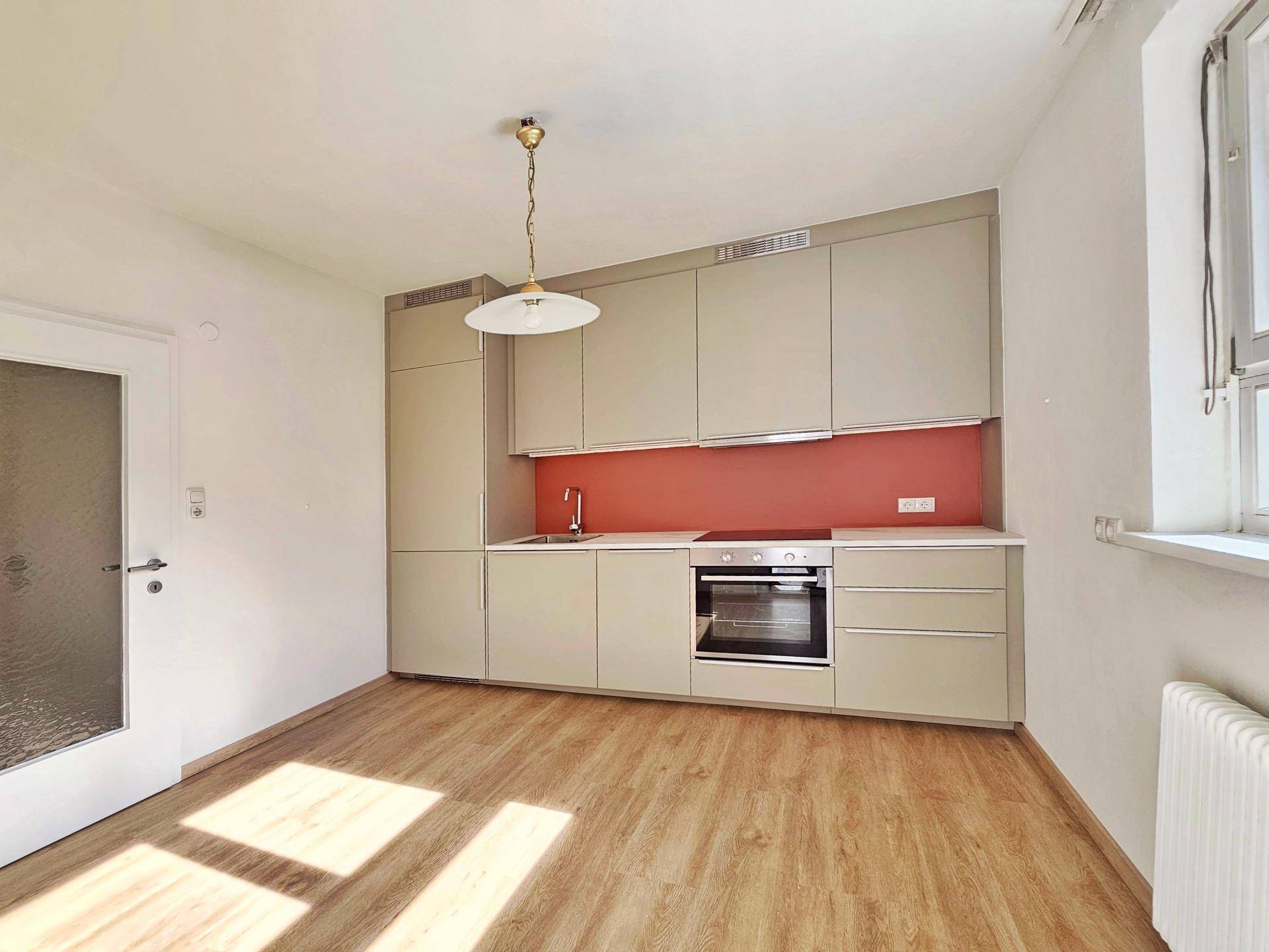 Moderne Einbauküche mit beigen Schränken, roter Rückwand und Holzoptikboden in einem hellen Raum.