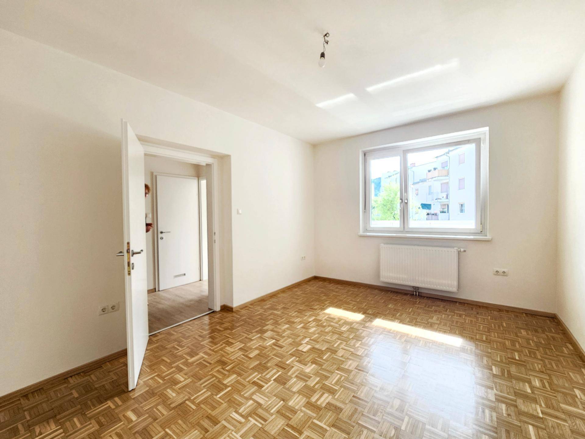 Helles, leeres Zimmer mit Parkettboden und großem Fenster, das viel Tageslicht hereinlässt.