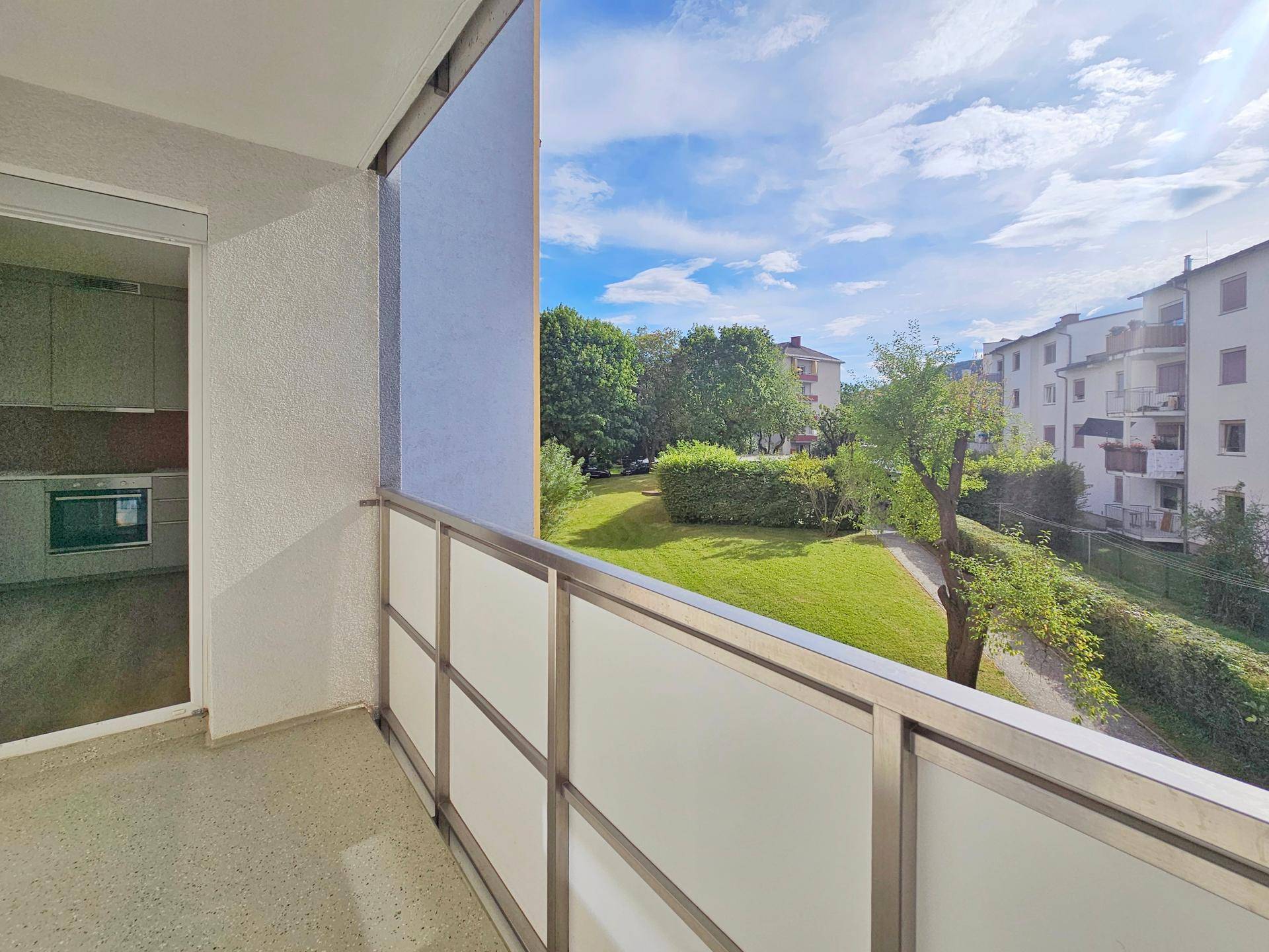 Balkon mit Metallgeländer und Blick auf eine grüne Gartenanlage und umliegende Gebäude.