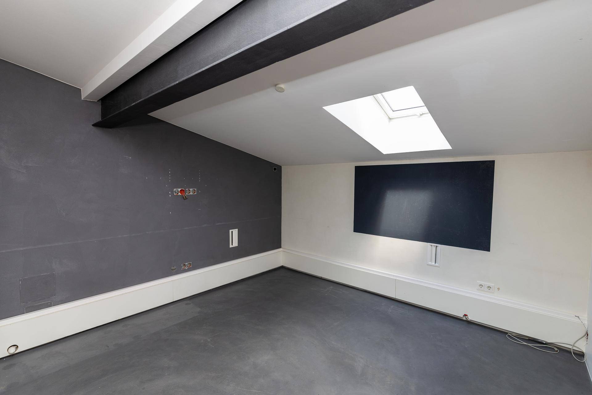 Moderner Raum mit grauen Wänden, Betonboden, Oberlicht und einem markanten schwarzen Deckenbalken.