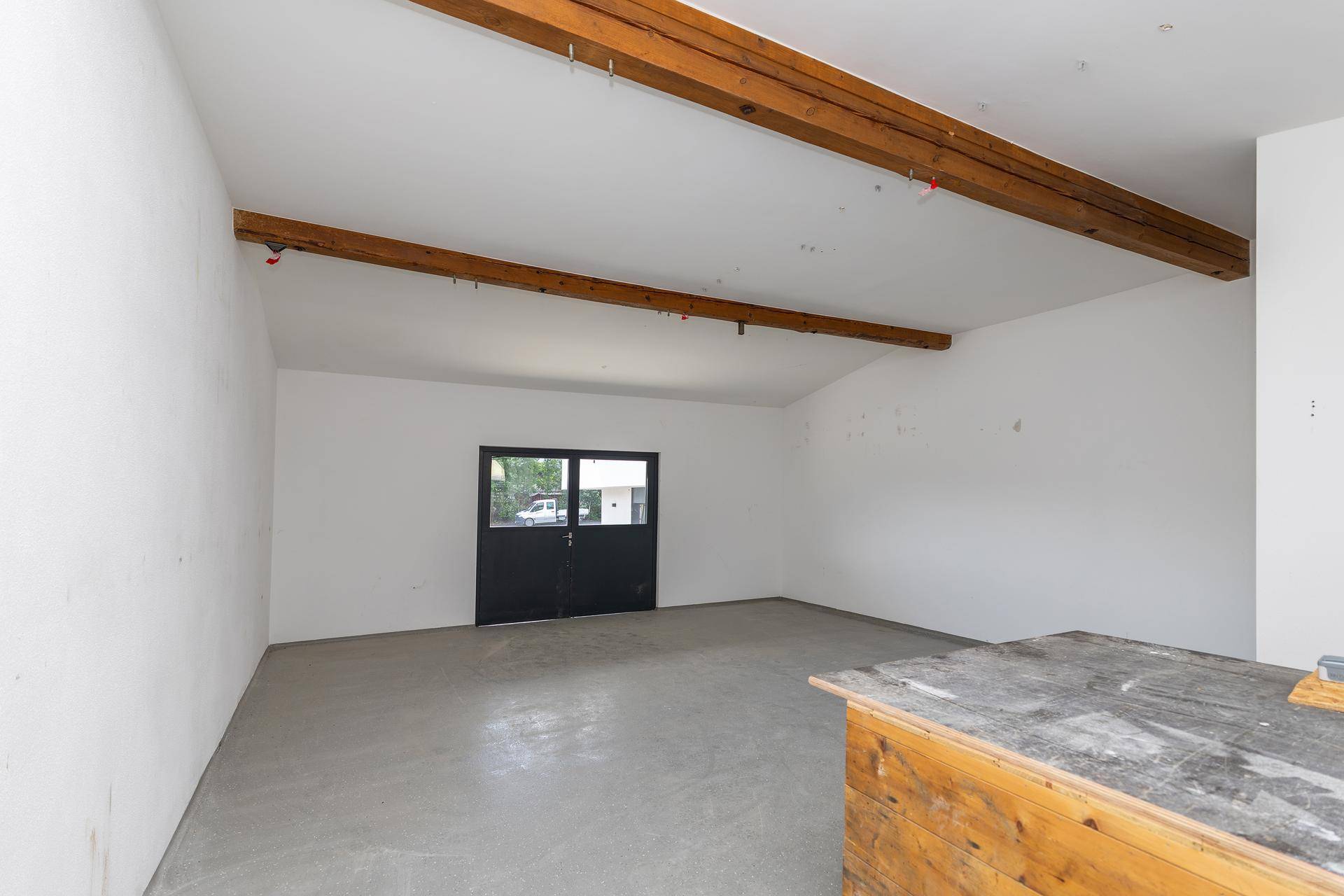 Leerer Raum mit Betonboden, weißen Wänden und sichtbaren Holzbalken, mit Zugang nach draußen.