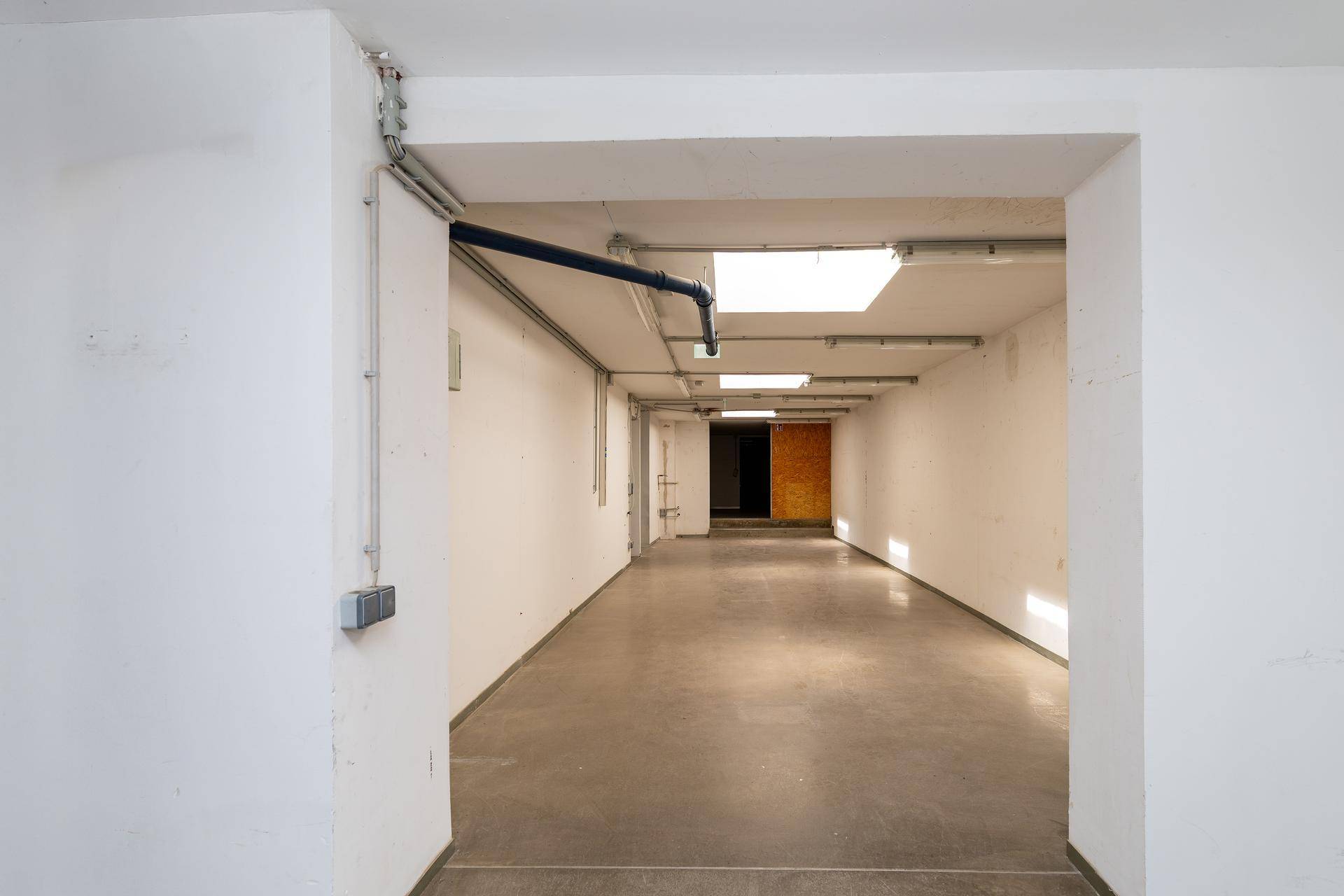 Langer, leerer Flur oder Lagerbereich mit Betonboden, sichtbaren Rohren und Oberlichtern für natürliches Licht.