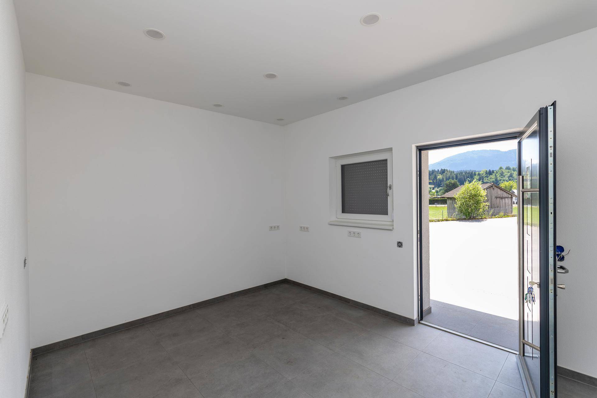 Heller Raum mit grauen Fliesen, weißen Wänden, Fenster mit Rollladen und offenem Zugang zur Natur.